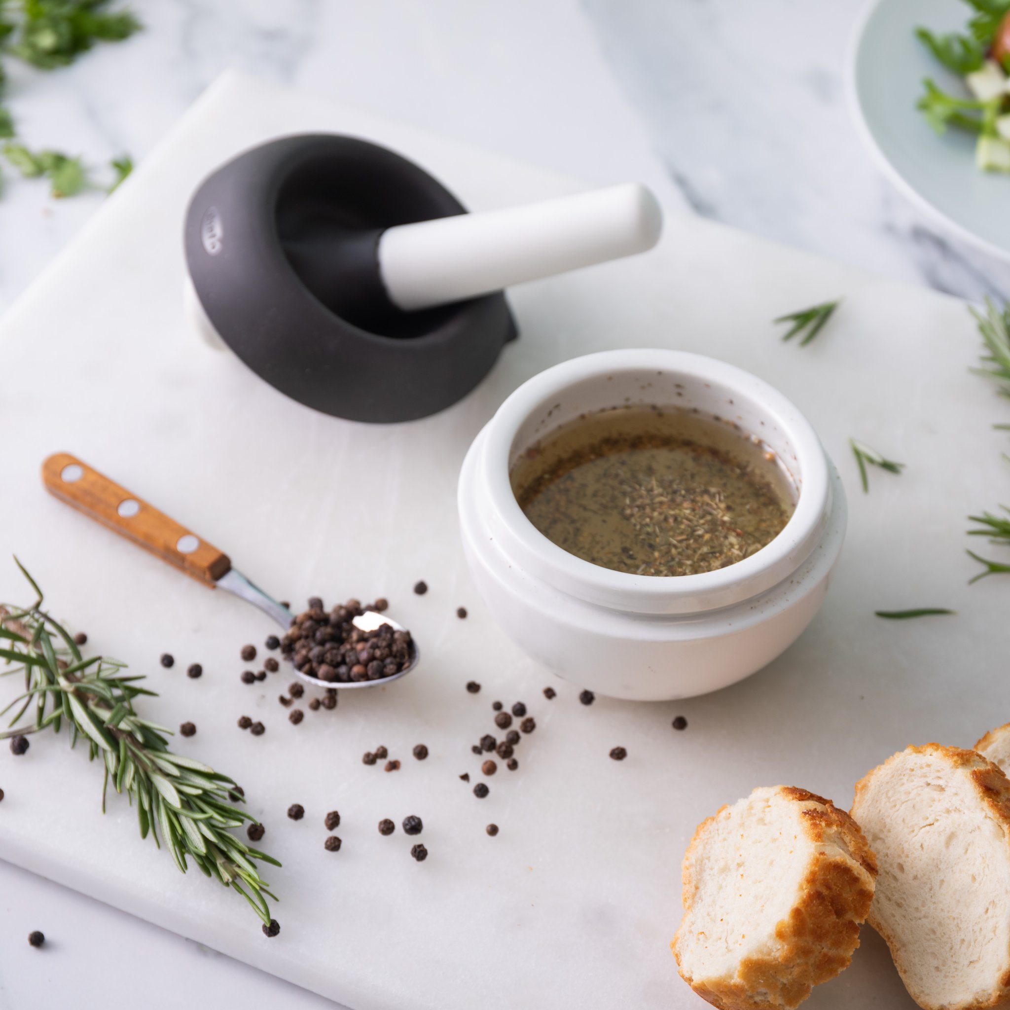 Keramikschale mit Pfeffer-Kräuter-Öl, daneben Spritzschutz mit Stössel und Brot
