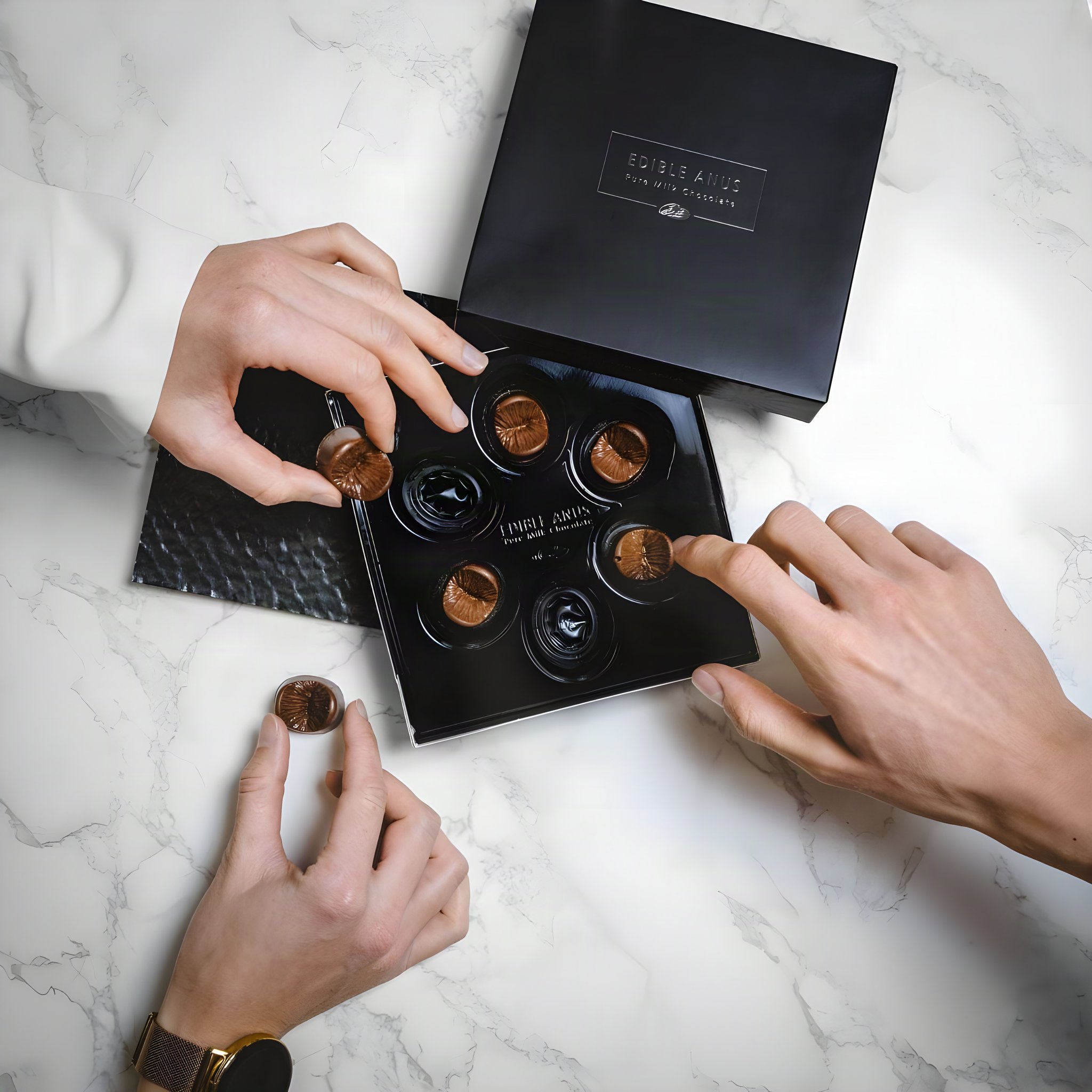 Mani che prendono cioccolatini a forma di ano dalla scatola nera su un piano di marmo