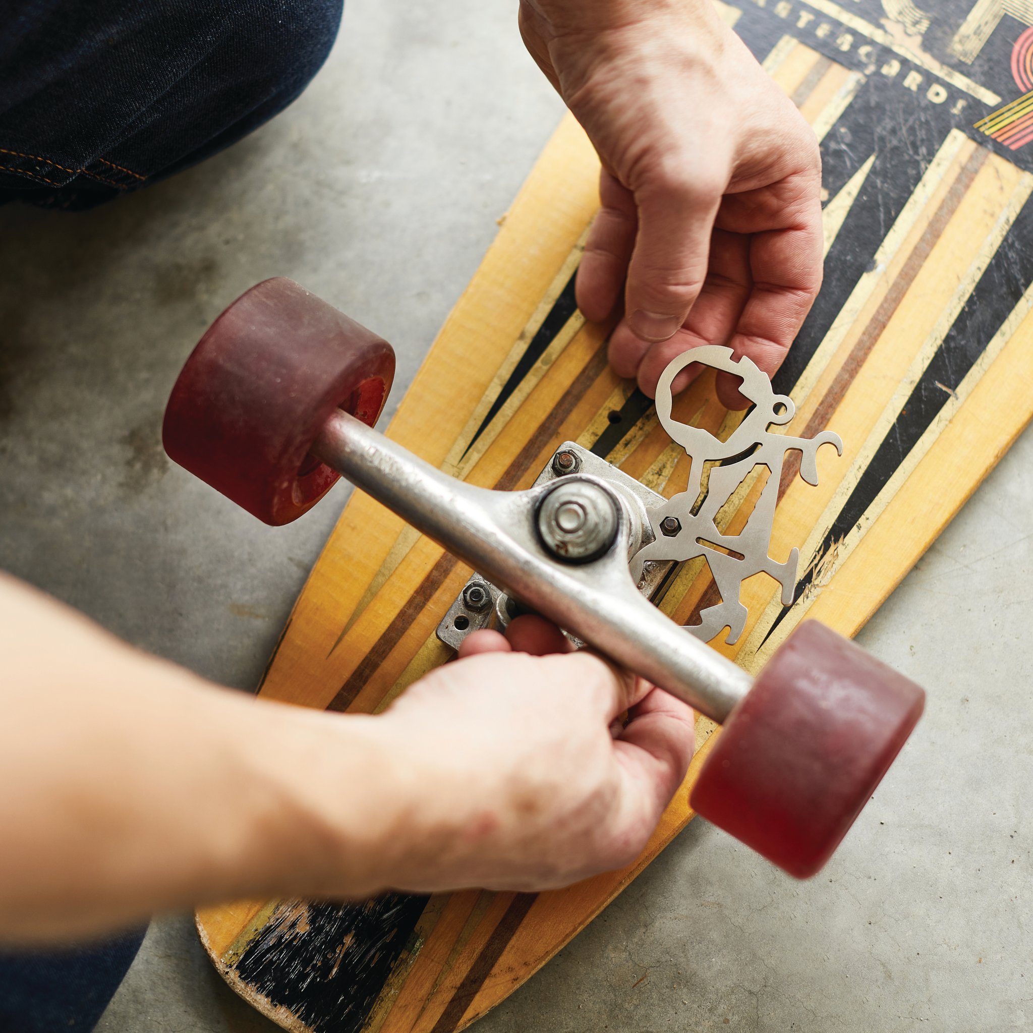 Hands holding the Mini Bicycle Multi-Tool against the axle of an upturned skateboard on concrete