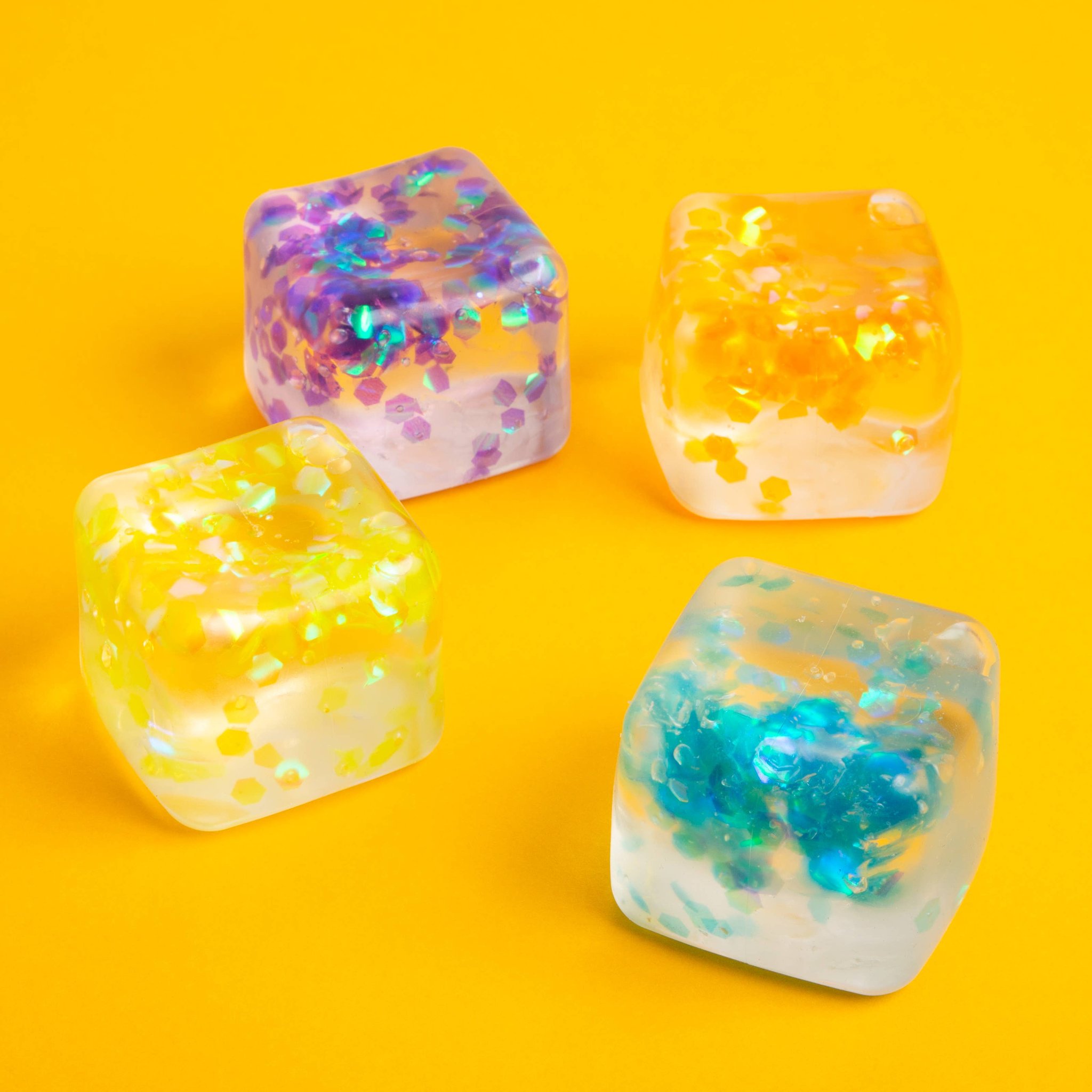 Vier Squishy Würfel mit Glitzerperlen im Inneren auf gelbem Hintergrund