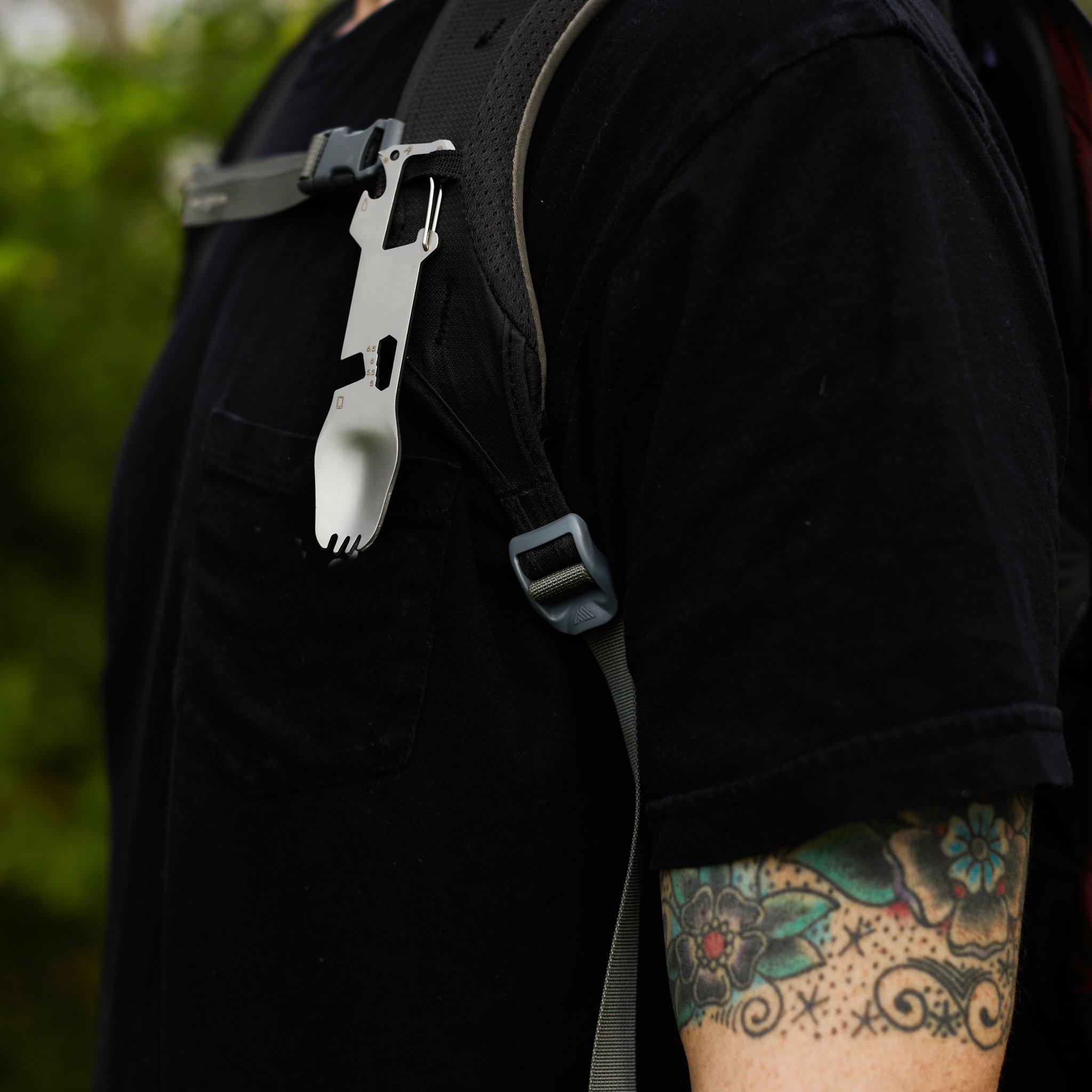 Vue latérale de la fourchette multifonctions sur une bretelle de sac, avec un bras tatoué et un fond de verdure flou