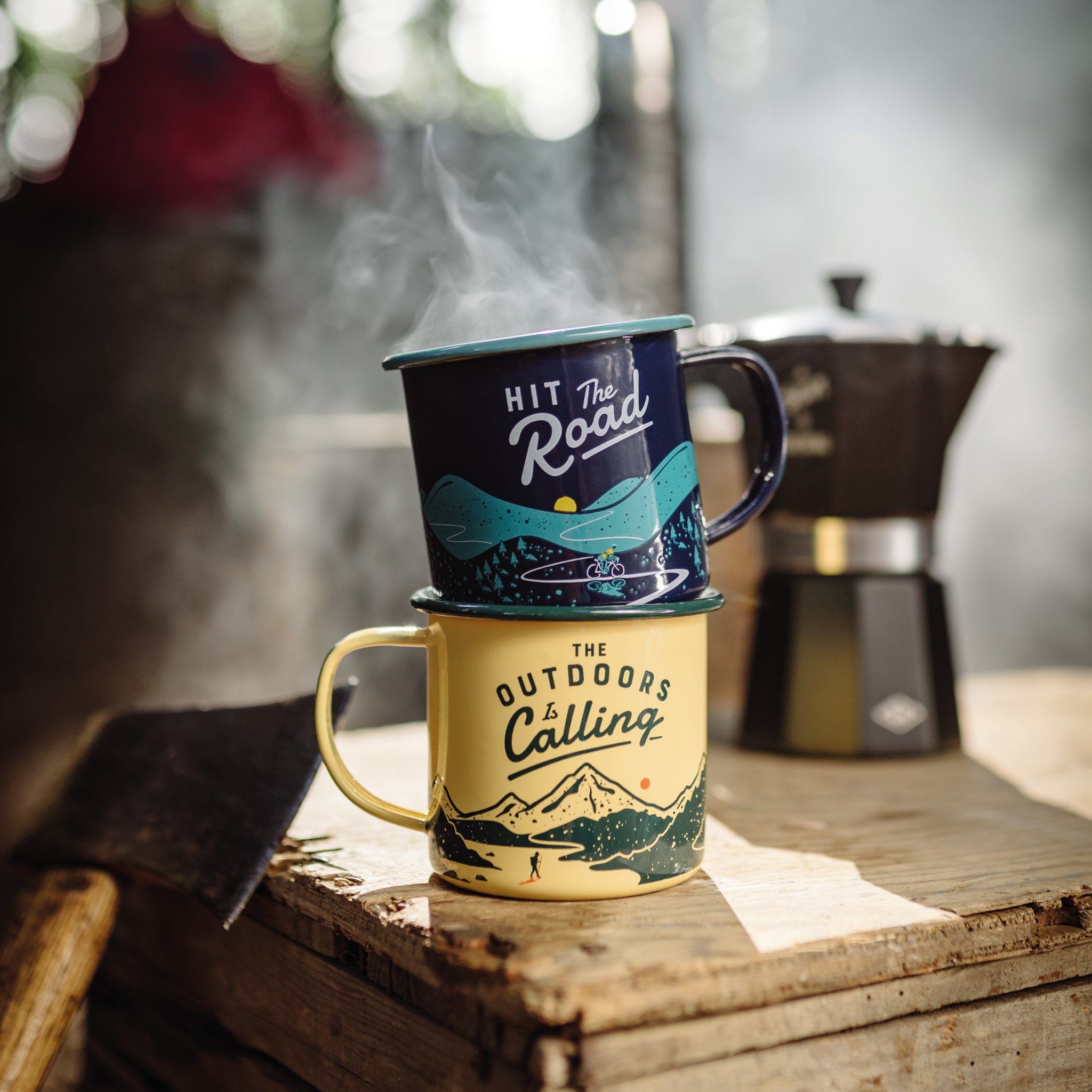 Dampfende Outdoor Emaille-Tasse, gestapelt unter einer blauen Tasse, auf einer Holzkiste neben Espressokocher und Axt