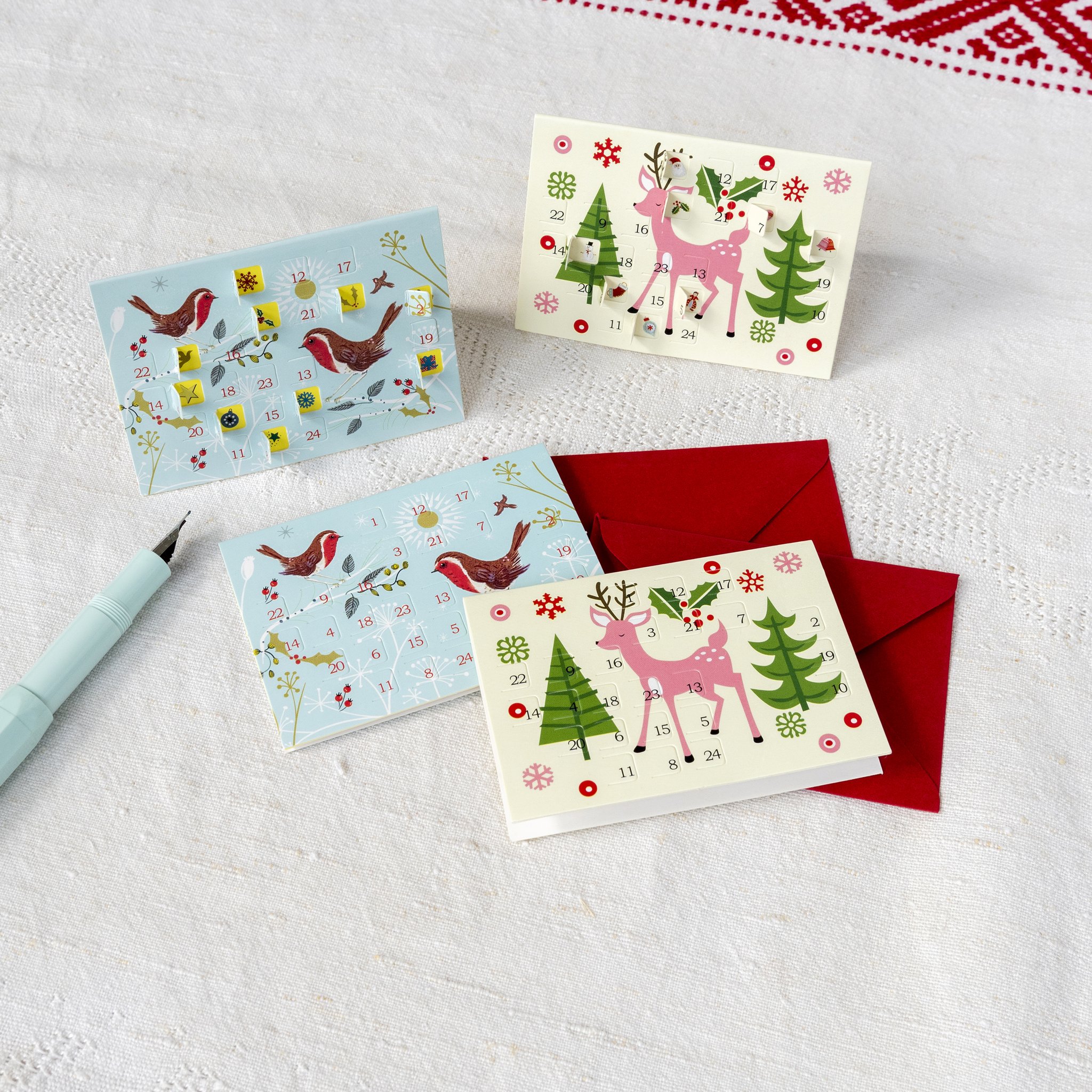 Weihnachtskarten-Adventskalender Rotkehlchen mit roten Couverts auf weissem Tuch, Stift daneben