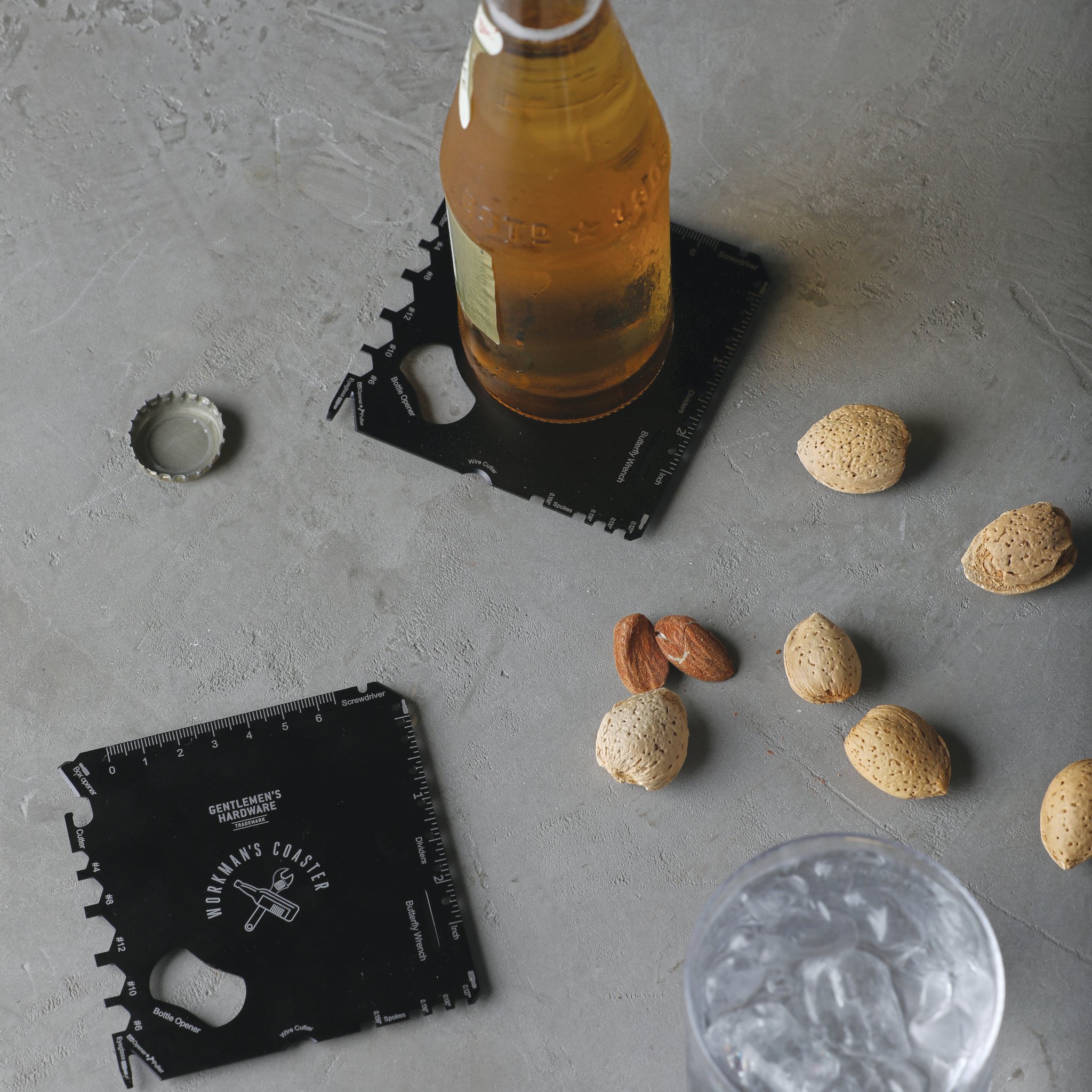 Su un tavolo di cemento una bottiglia di birra poggia su un sottobicchiere multiutensile, con tappo, mandorle e bicchiere