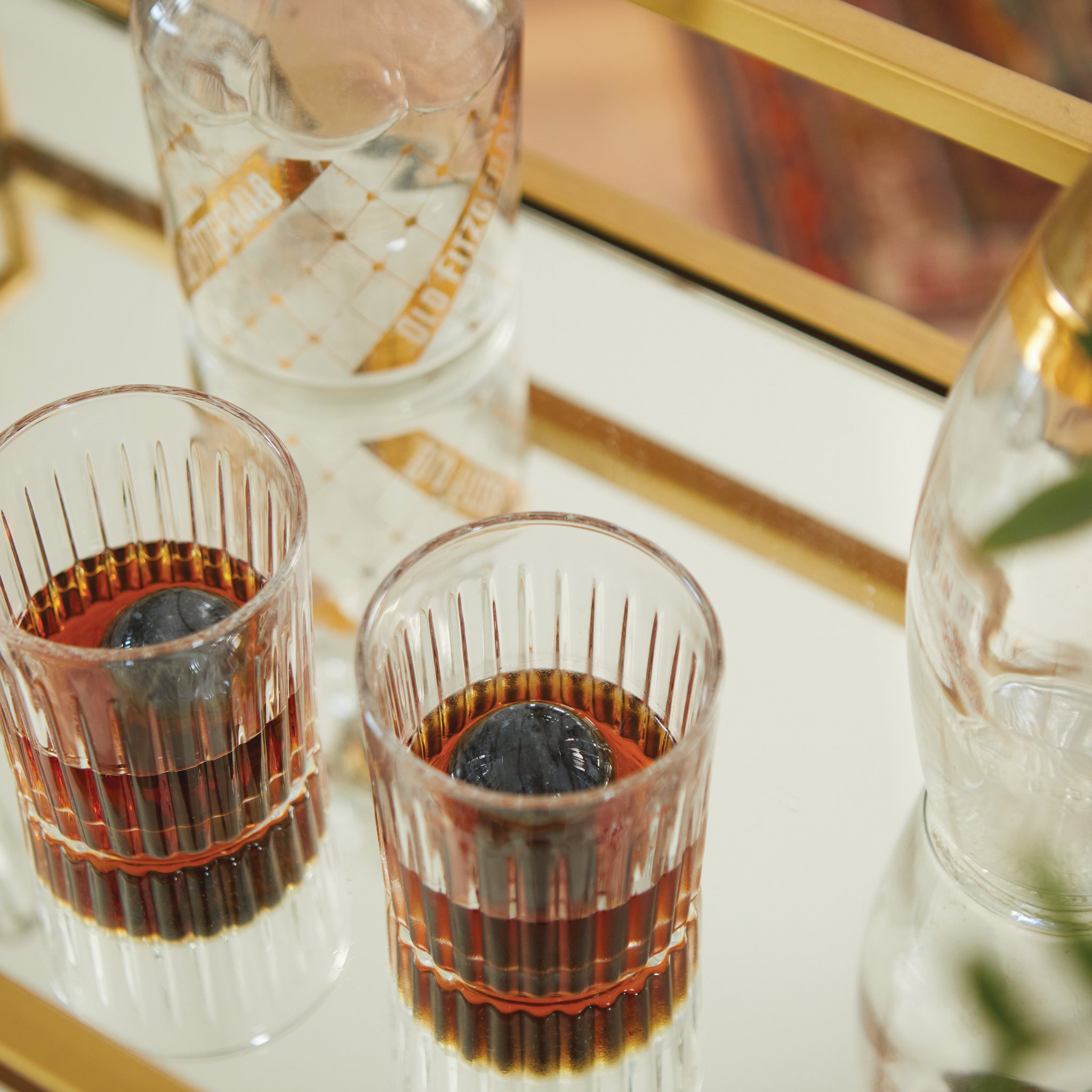 Zwei Gläser mit Whisky und Granitkugel auf einem Spiegeltablett mit Goldrahmen, dahinter eine Karaffe