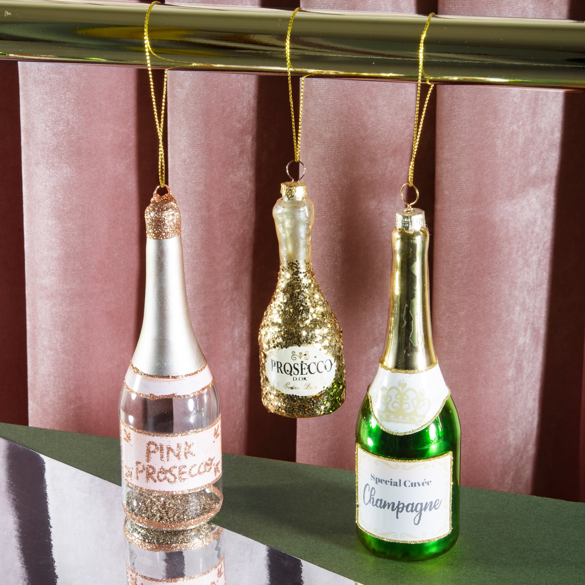 Décoration de sapin bouteille de prosecco suspendue parmi des ornements champagne et rosé devant du velours rose