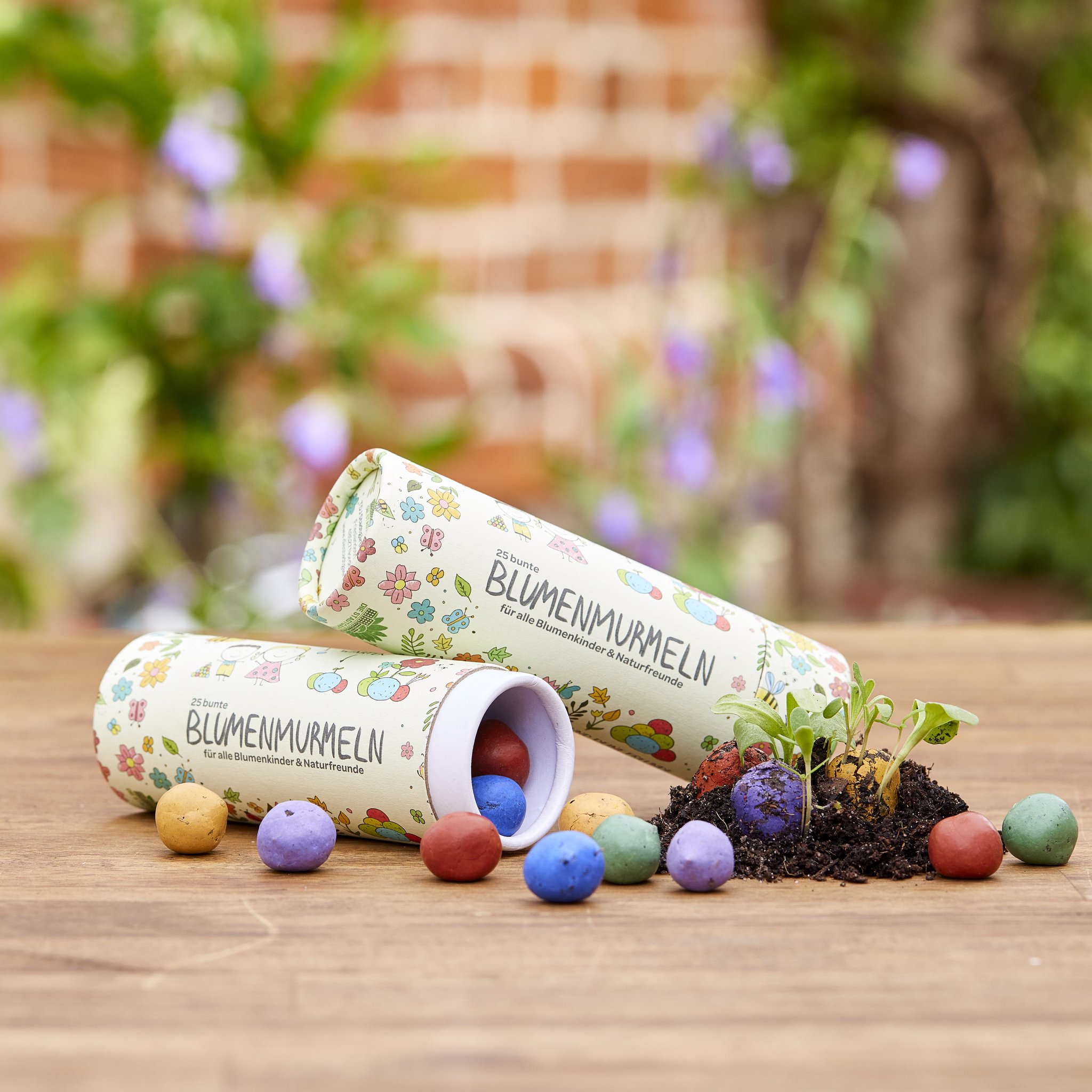 Zwei Dosen Blumenmurmeln auf einem Holztisch, davor bunte Samenkugeln und Erde mit Keimlingen