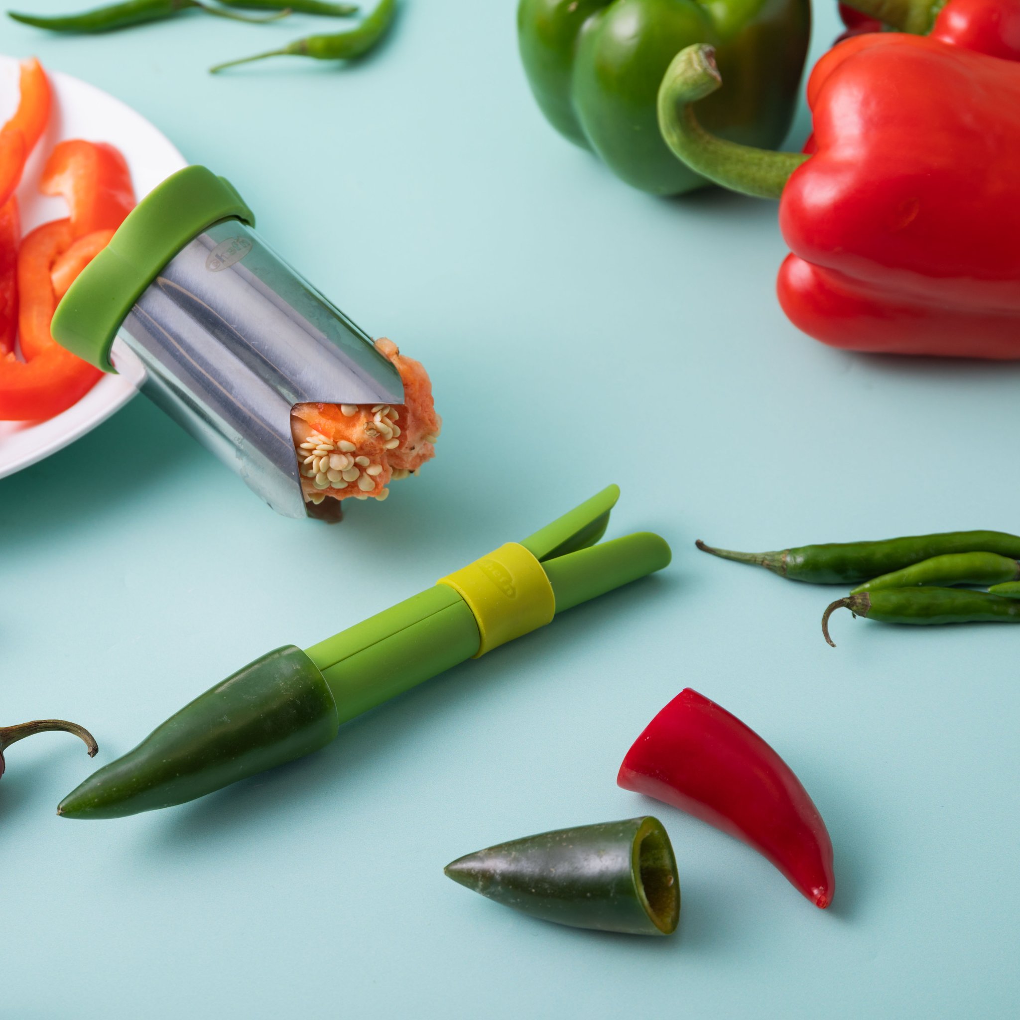 Entkerner mit Paprikakernen neben Jalapeños und Paprikastreifen auf türkisem Grund