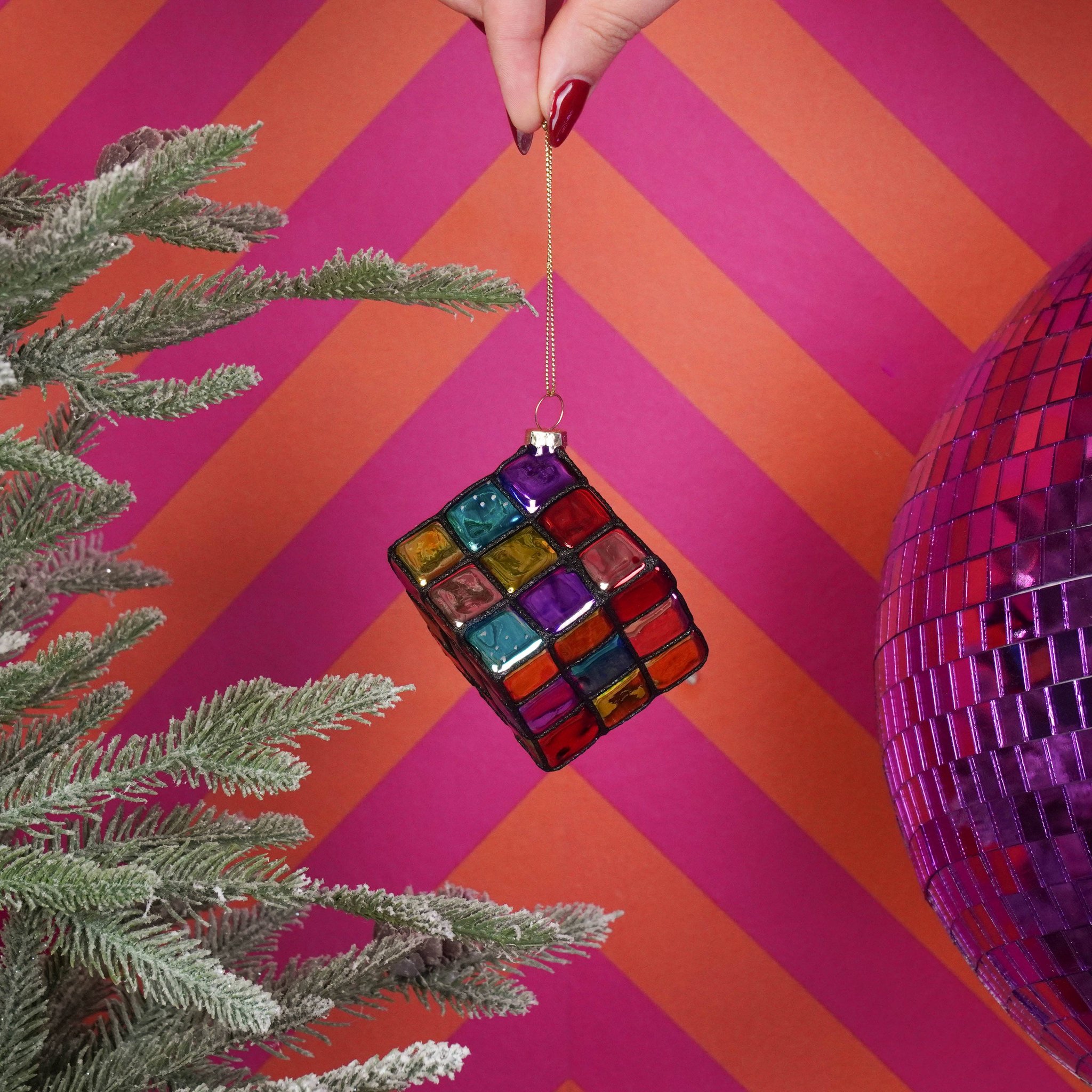 Bunter Zauberwürfel Weihnachtsanhänger aus Glas vor pinkem Chevron-Hintergrund mit Discokugel