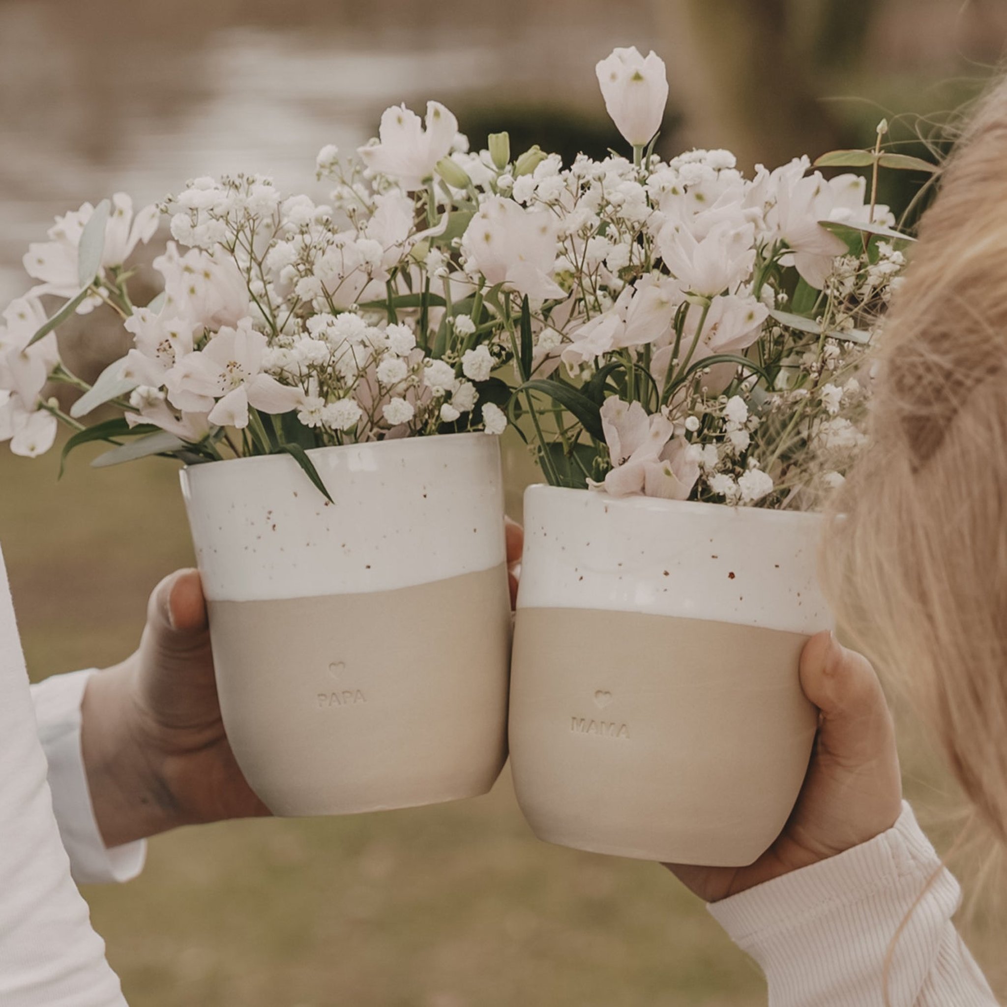 Zwei Becher Mama mit Schleierkraut und weissen Blumen draussen in Kinderhänden