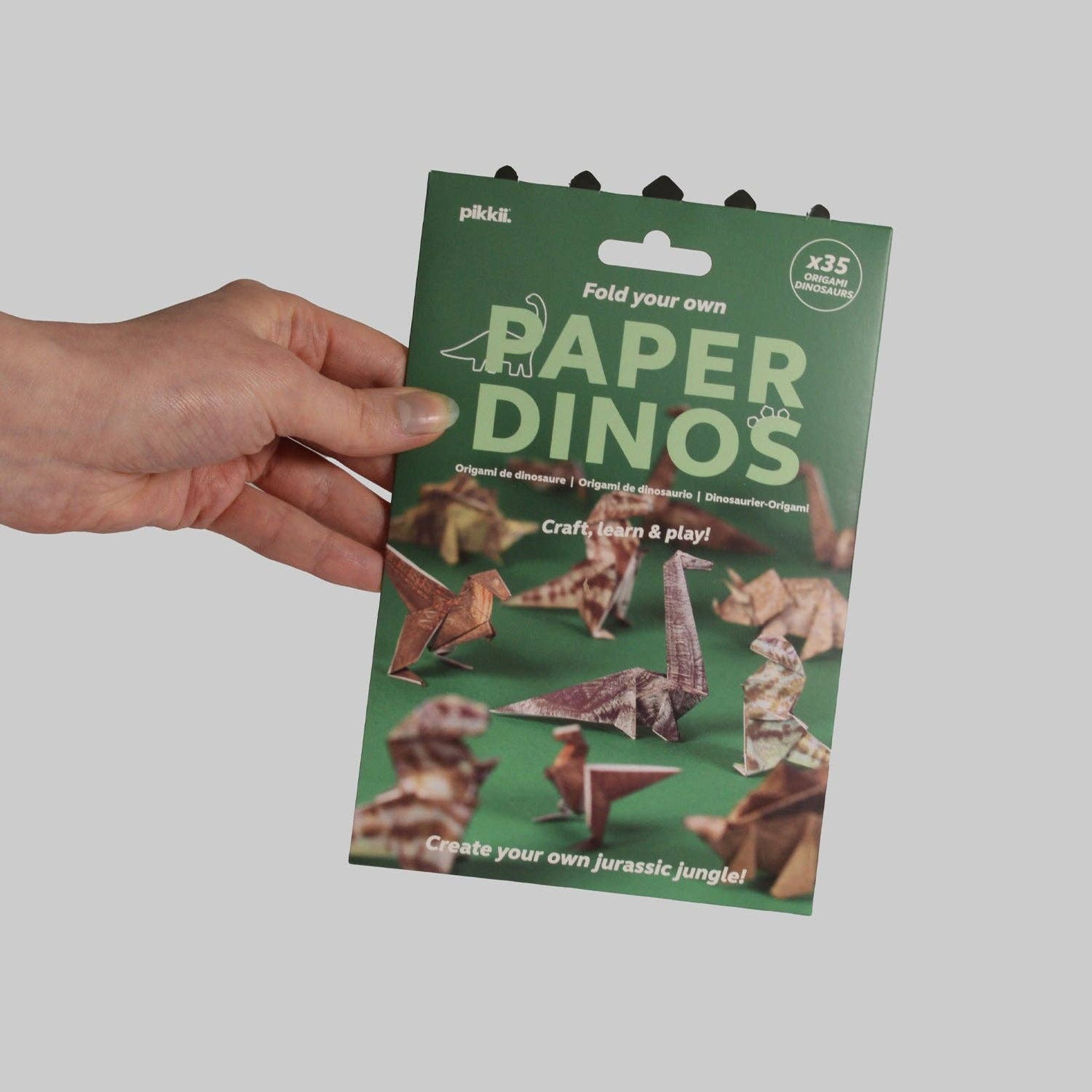 Hand holding the pack of the Pikkii Paper Dinos origami kit