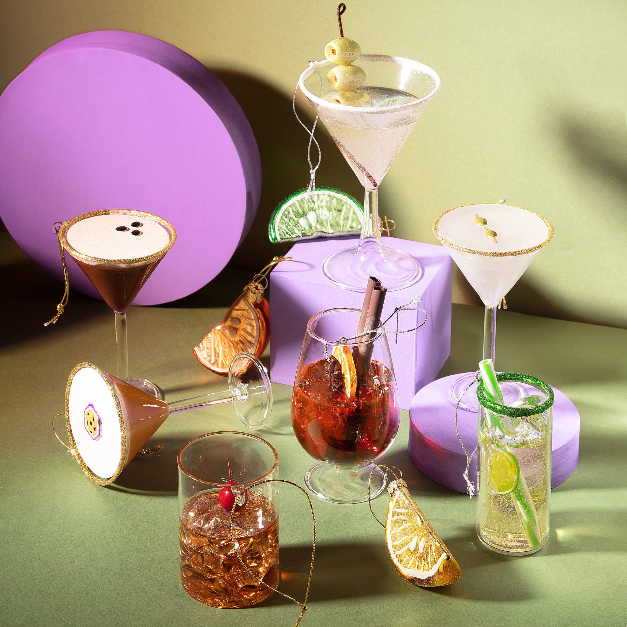 Whiskyglas Weihnachtsbaumschmuck von Sass & Belle zwischen Cocktail-Anhängern auf lila Podesten