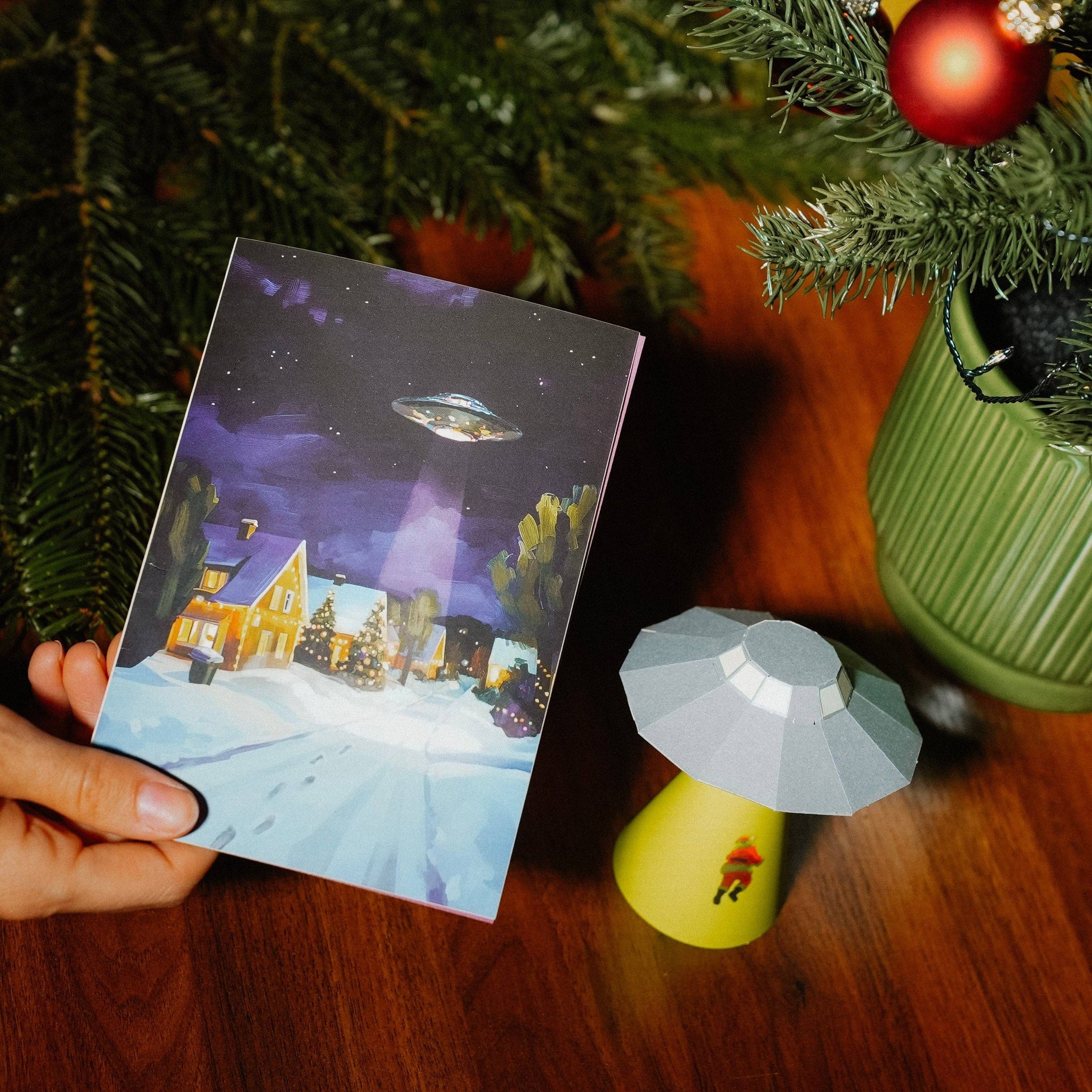 Hand hält die Weihnachtskarte mit UFO über verschneitem Dorf neben dem gebastelten 3D-Modell
