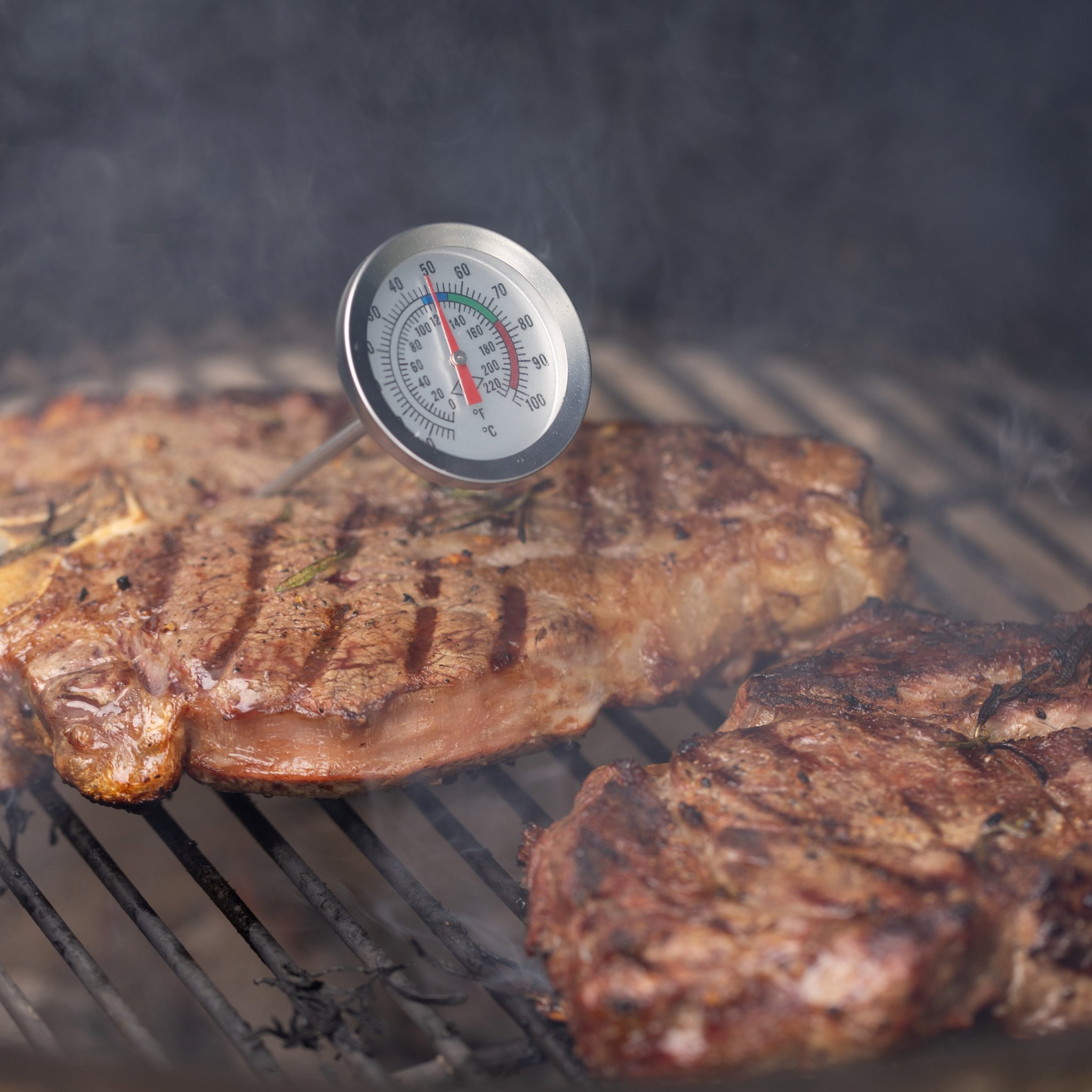 Angled view of the thermometer in a steak on the barbecue, its needle in the red zone, smoke in the dark background