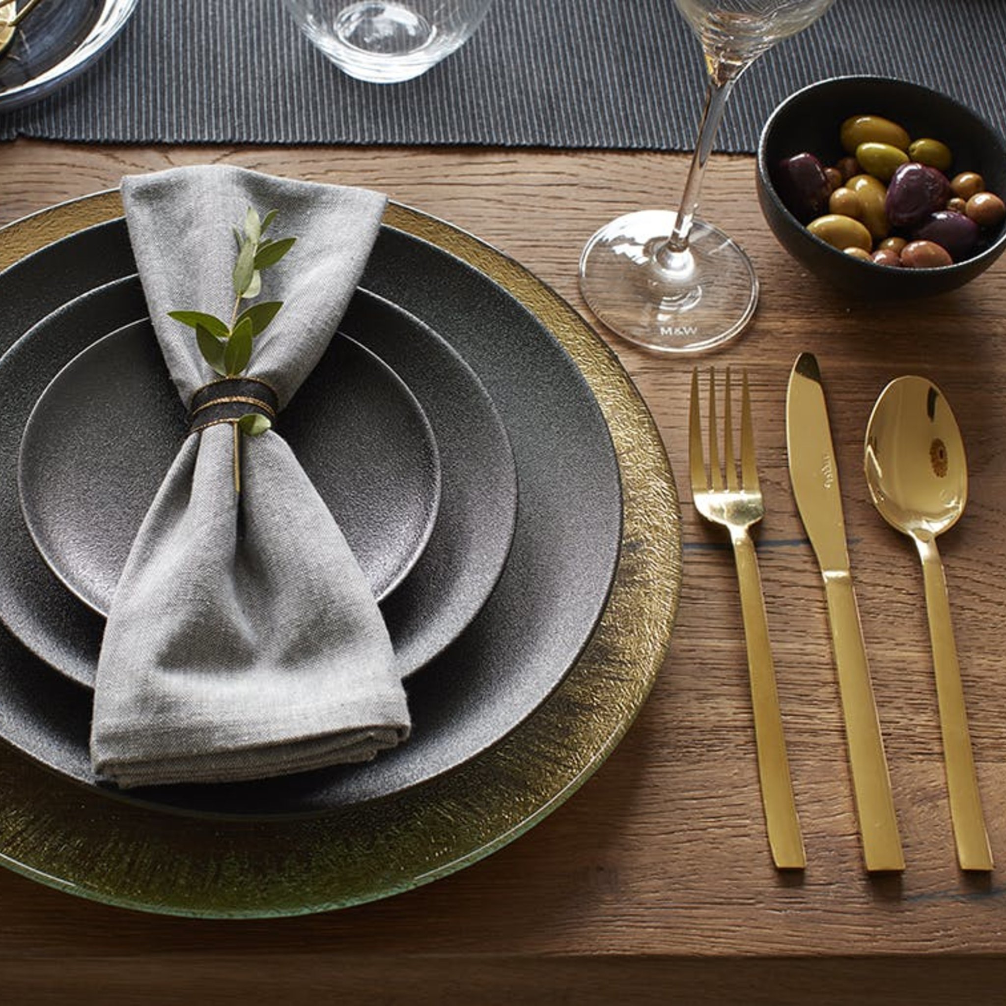 Gedeckter Holztisch mit dunklen Tellern, Serviette und goldenem Besteck daneben