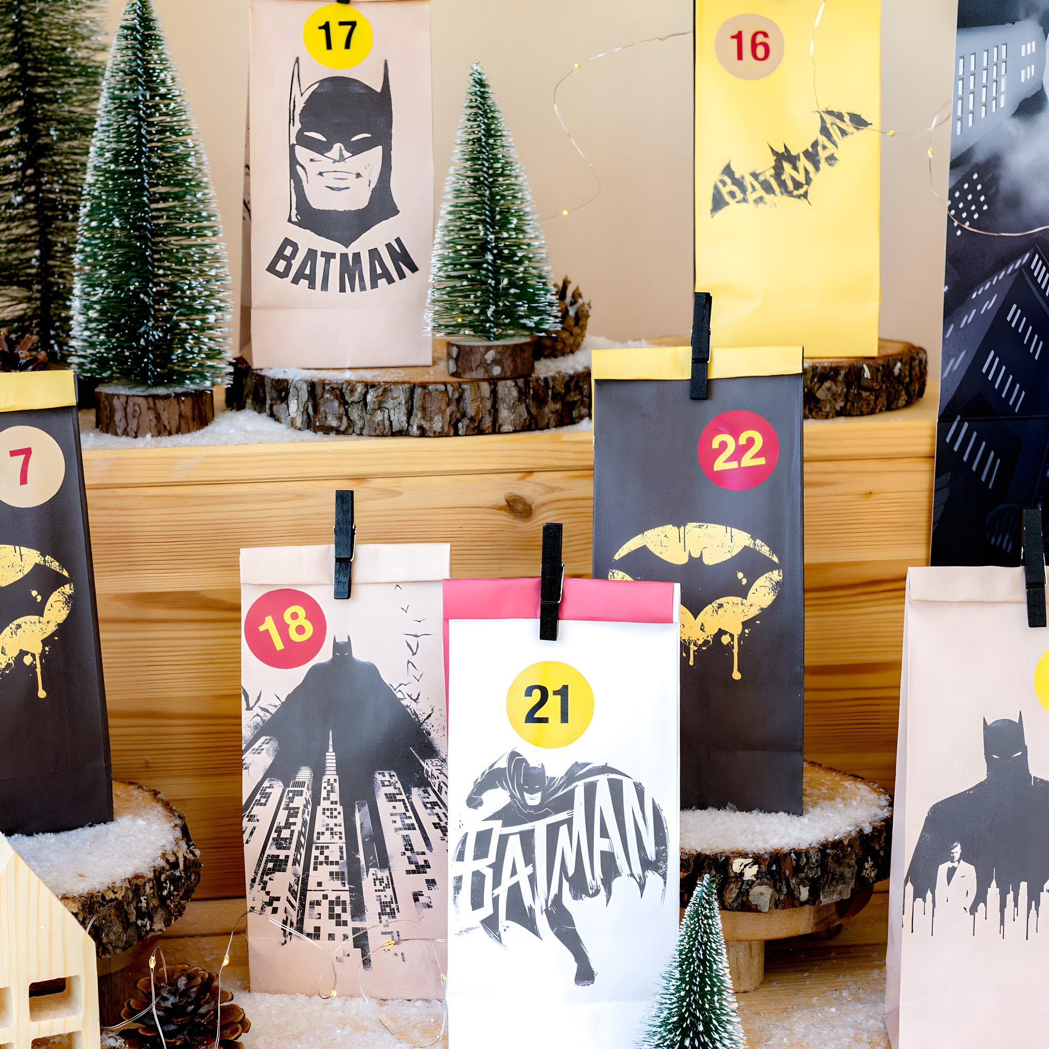 Batman Adventskalender-Tüten in Gelb, Schwarz, Pink und Kraftpapier mit Klammern auf Holzstufen zwischen Tannen