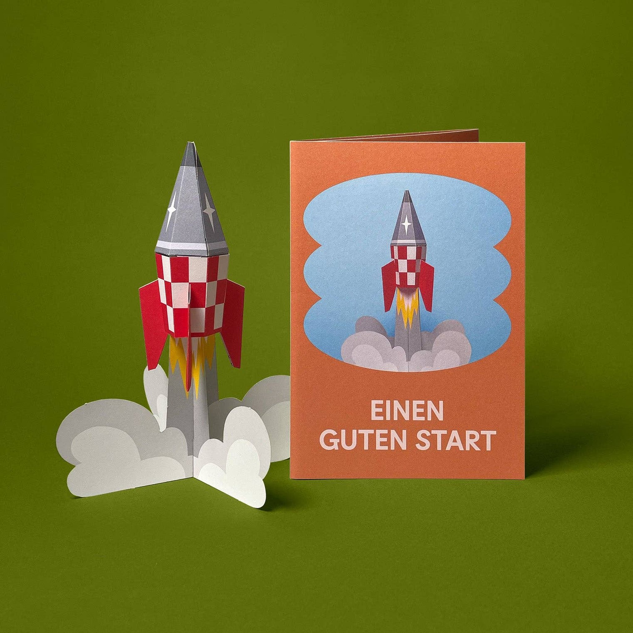 Gefaltetes 3D-Modell Retro-Rakete neben der Grusskarte «Einen guten Start» von Faltmeister