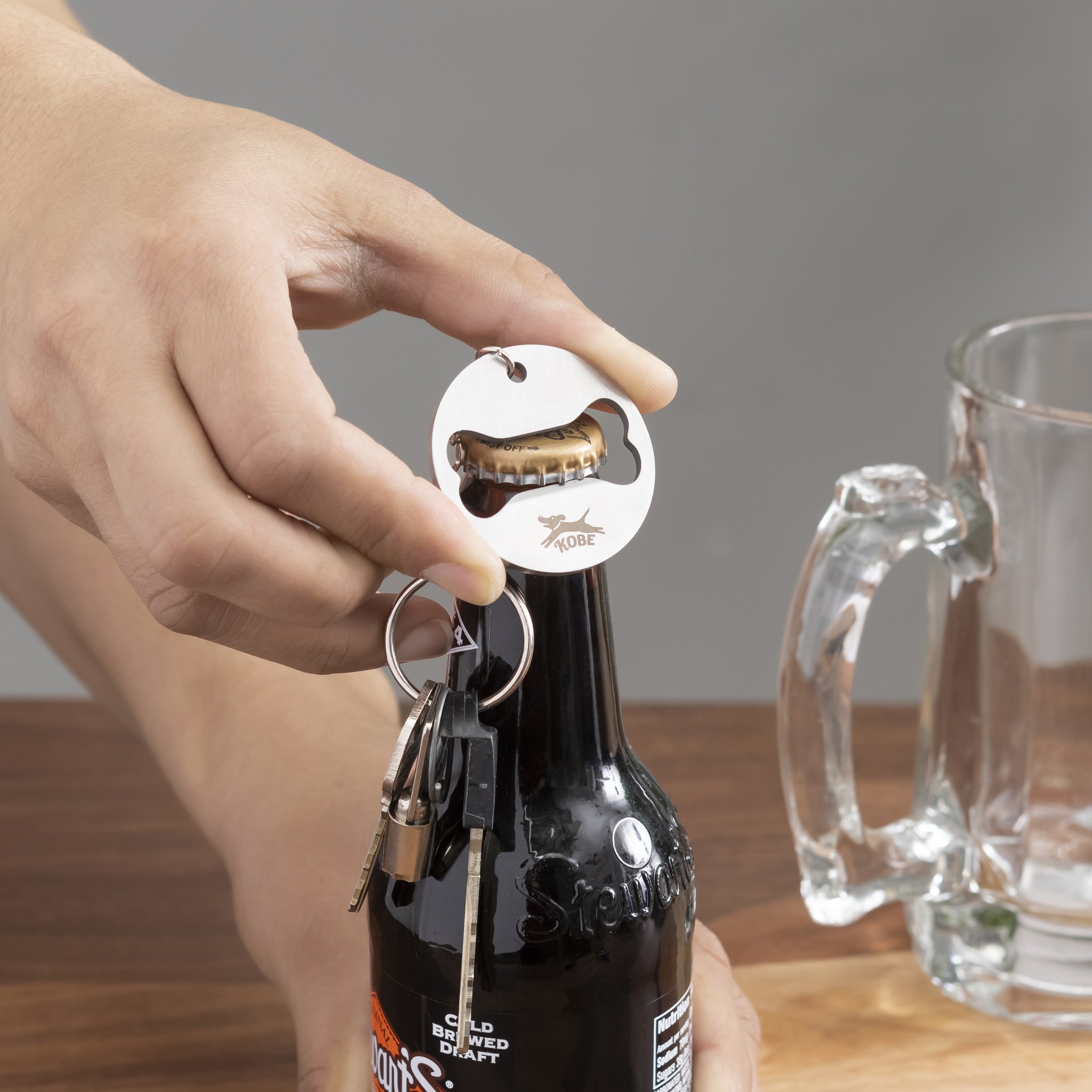Hand öffnet mit dem Anhänger eine Bierflasche neben einem Glas