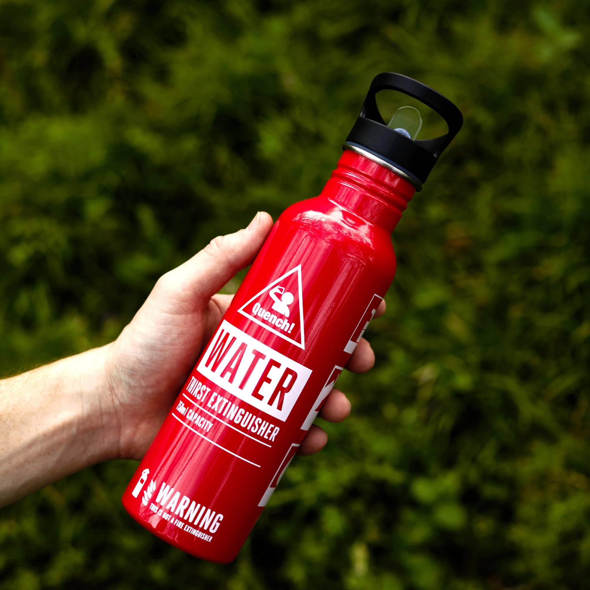 Bouteille rouge de 750 ml façon extincteur avec poignée tenue en main sur fond de verdure
