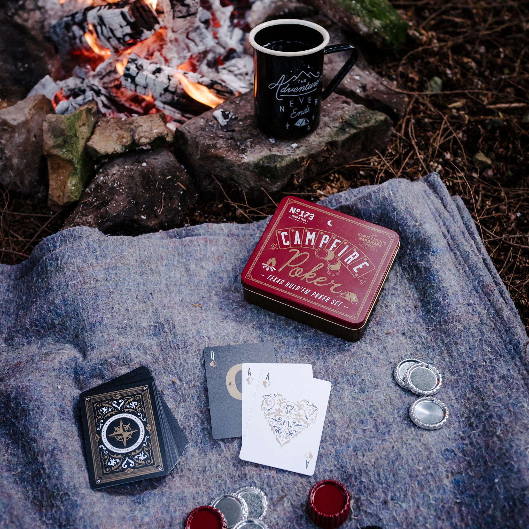 Campfire Poker tin, cards and chips on a grey blanket beside a campfire, with a black enamel mug resting on a stone