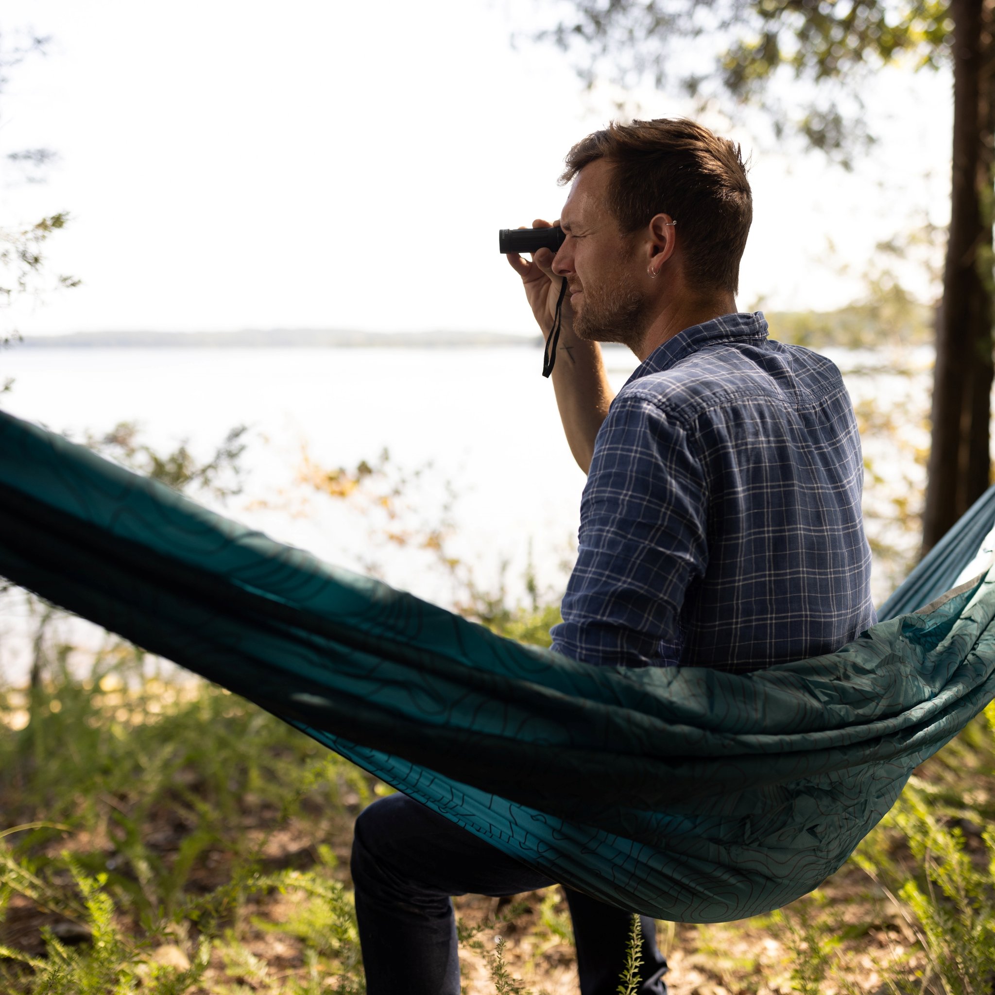Homme dans un hamac, le monoculaire à l'œil, avec arbres et lac calme derrière, Kit d'exploration en plein air