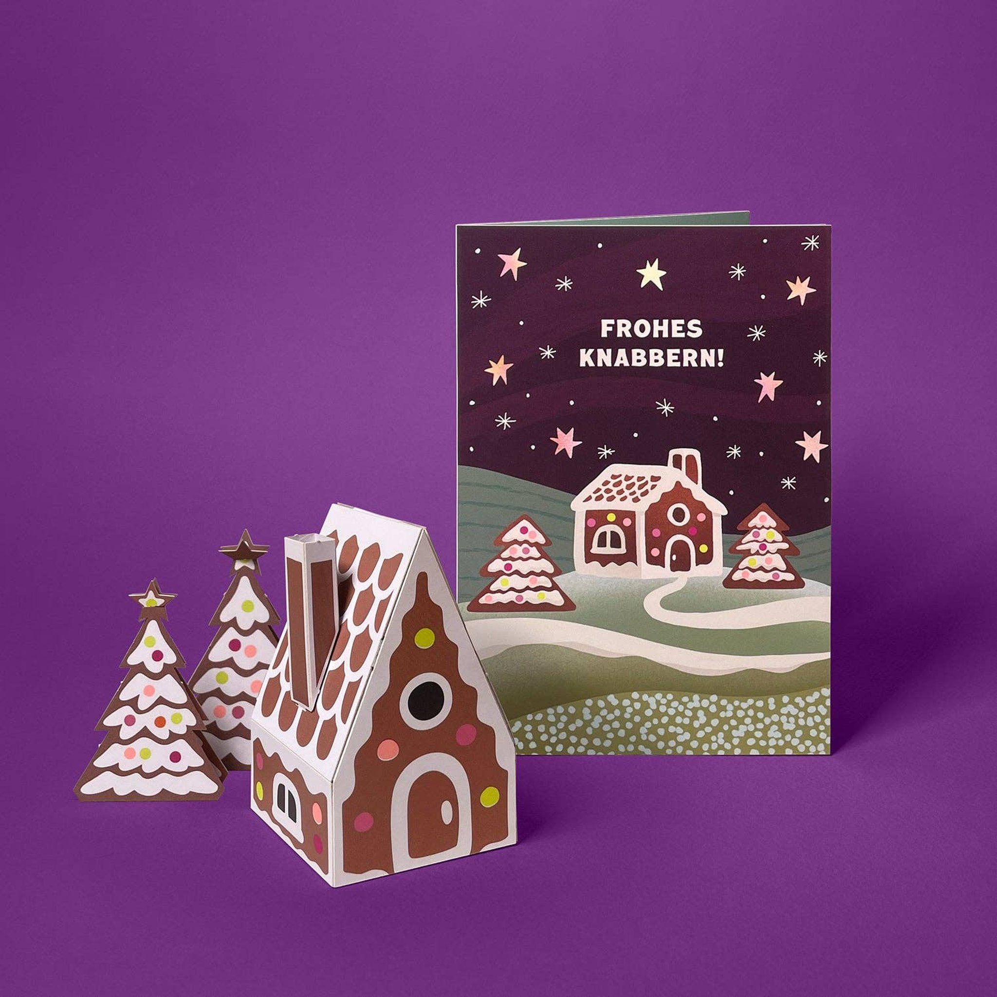 Gefaltetes 3D-Modell Lebkuchenhaus mit Tannenbäumen neben der Weihnachtskarte «Frohes Knabbern!» von Faltmeister