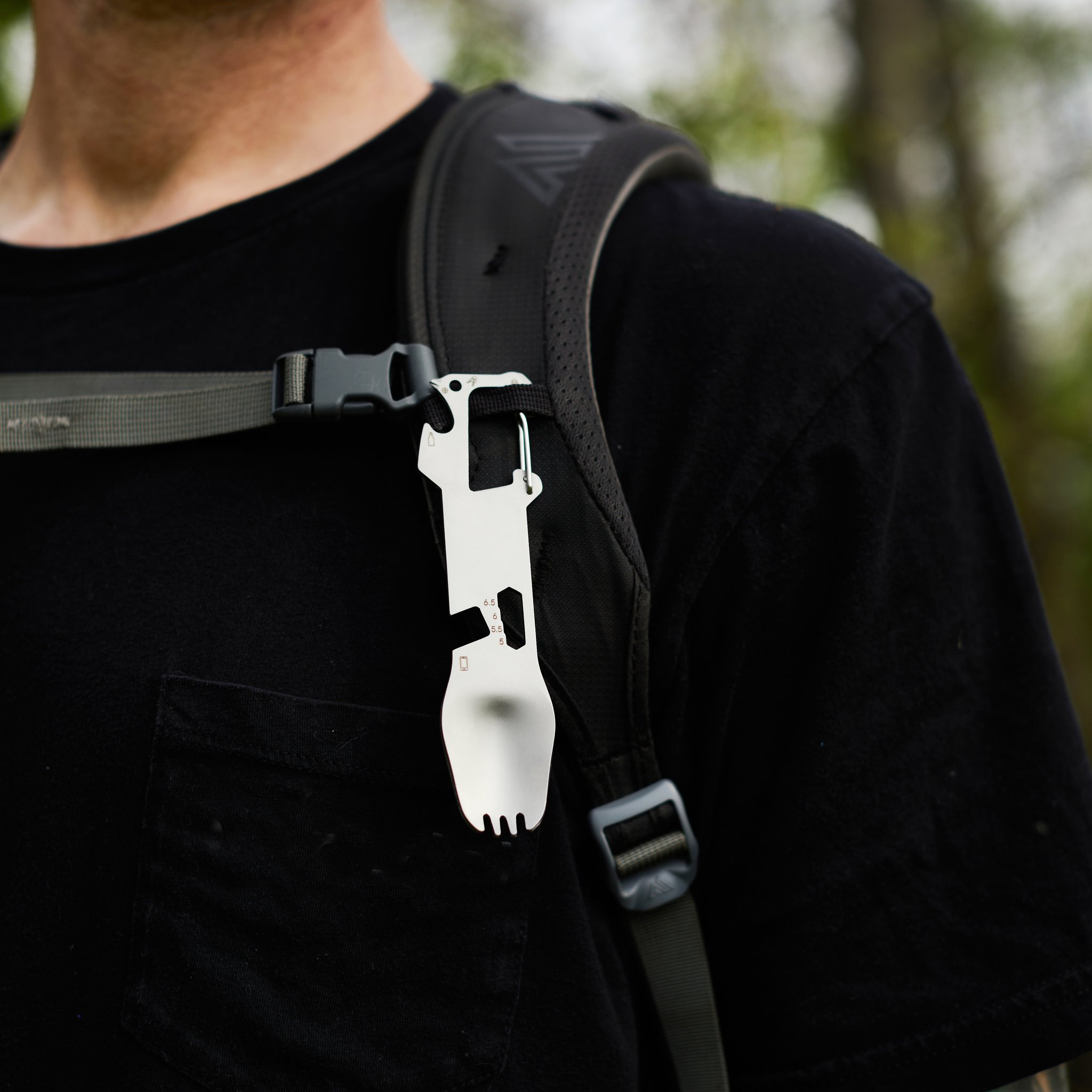 Fourchette multifonctions accrochée par son mousqueton à la bretelle d'un sac à dos, personne en t-shirt noir en forêt