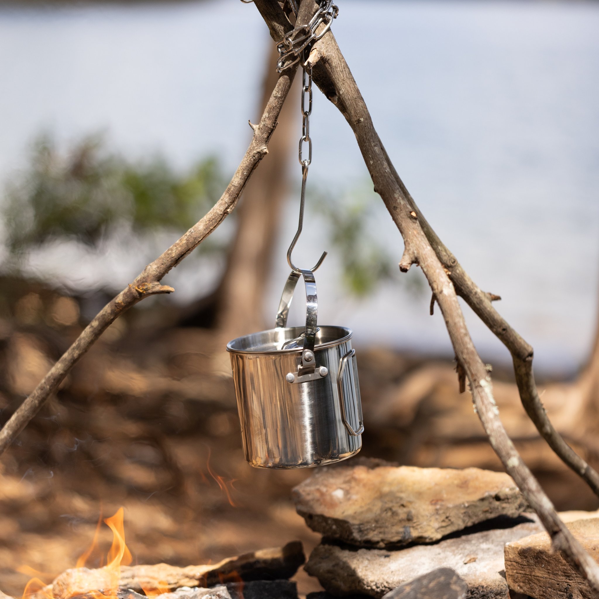 Campfire Cooking Pot on a chain from the branch tripod, with stones and small flames below and a blurred background