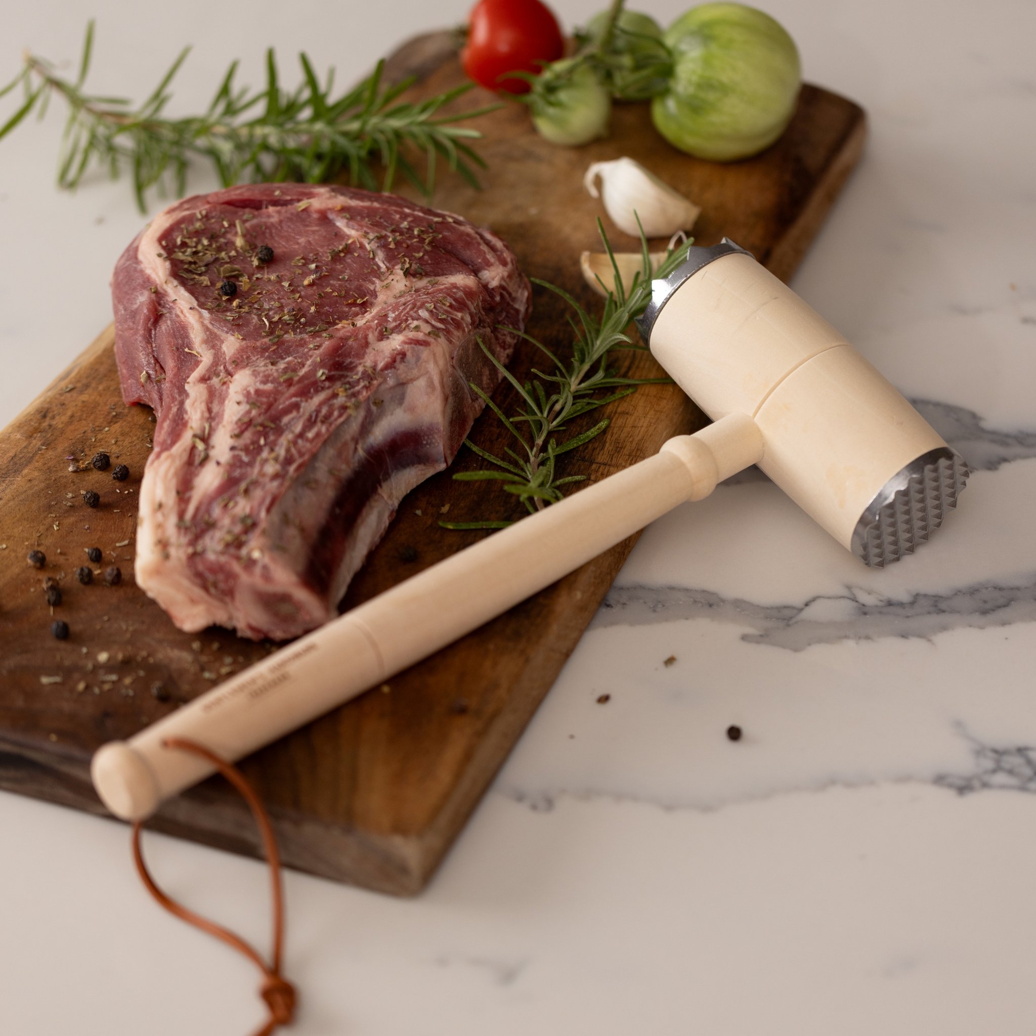 Meat tenderizer from the BBQ Lovers Kit on a wooden board beside a raw steak, rosemary, garlic and peppercorns