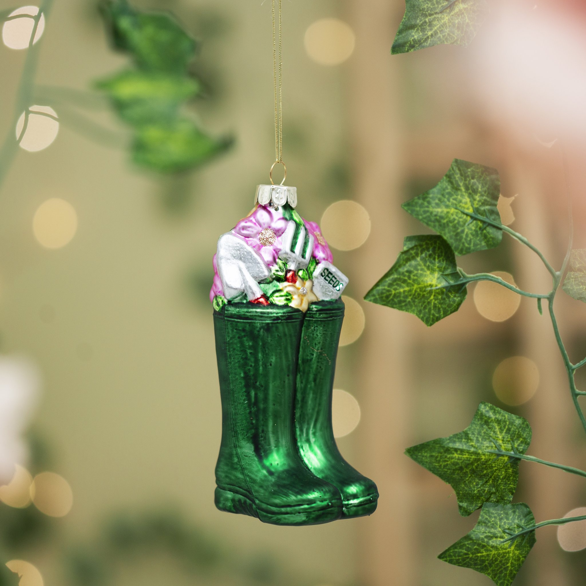 Sass & Belle Gummistiefel-Weihnachtsbaumschmuck zwischen Efeuranken mit warmem Bokeh-Lichtspiel – Lifestyle