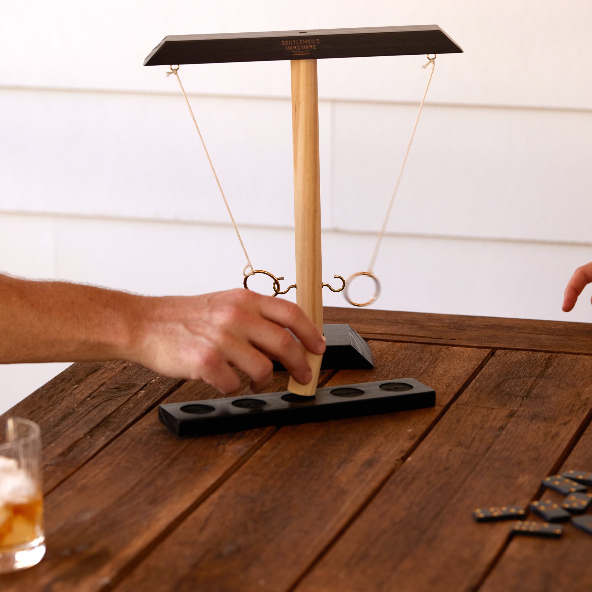 Sur une table en bois, une main saisit le plot du Jeu de Lancer d'Anneau, avec un verre et des dominos
