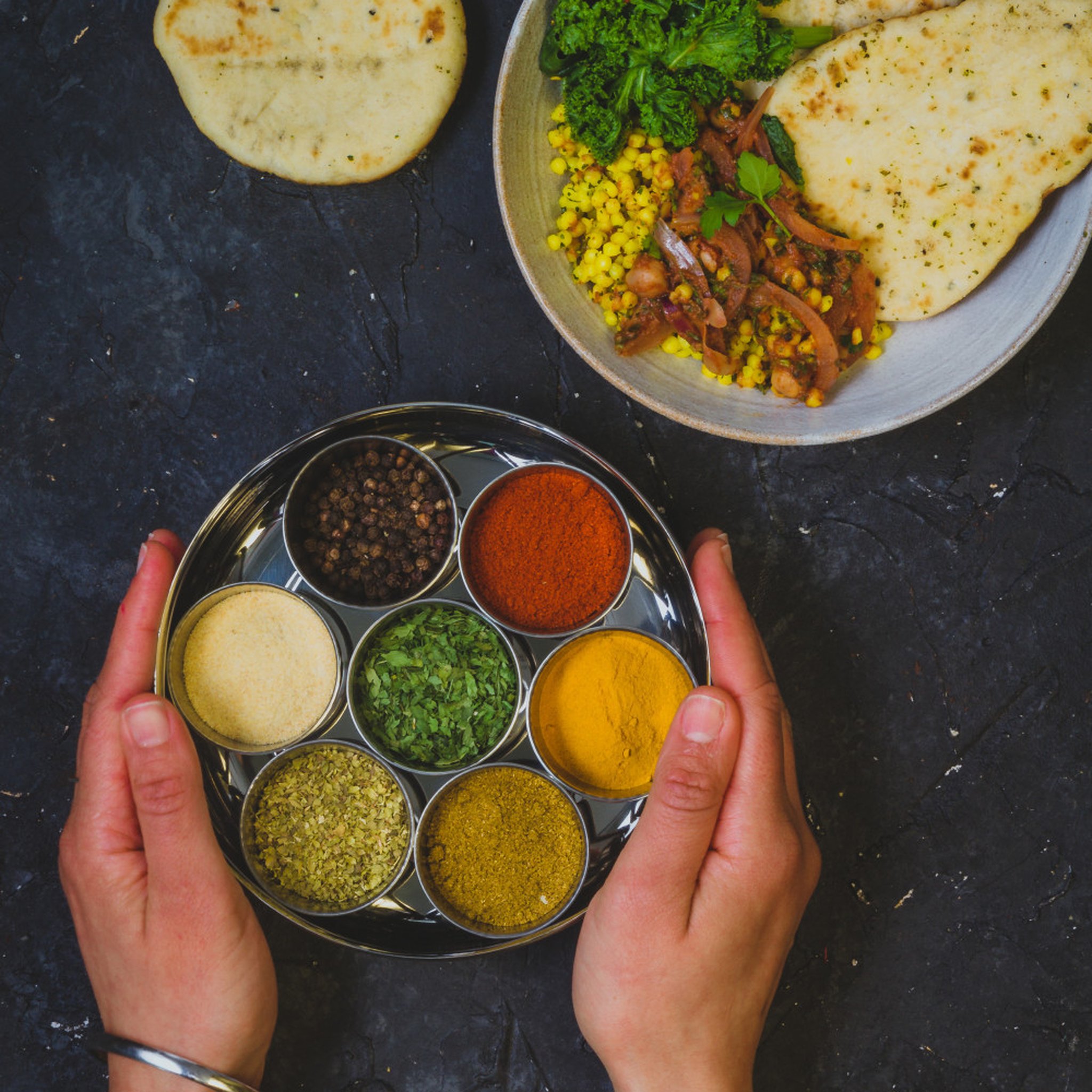Masala Dabba mit 7 Gewürzen gehalten von Händen, Naan und Curry im Hintergrund