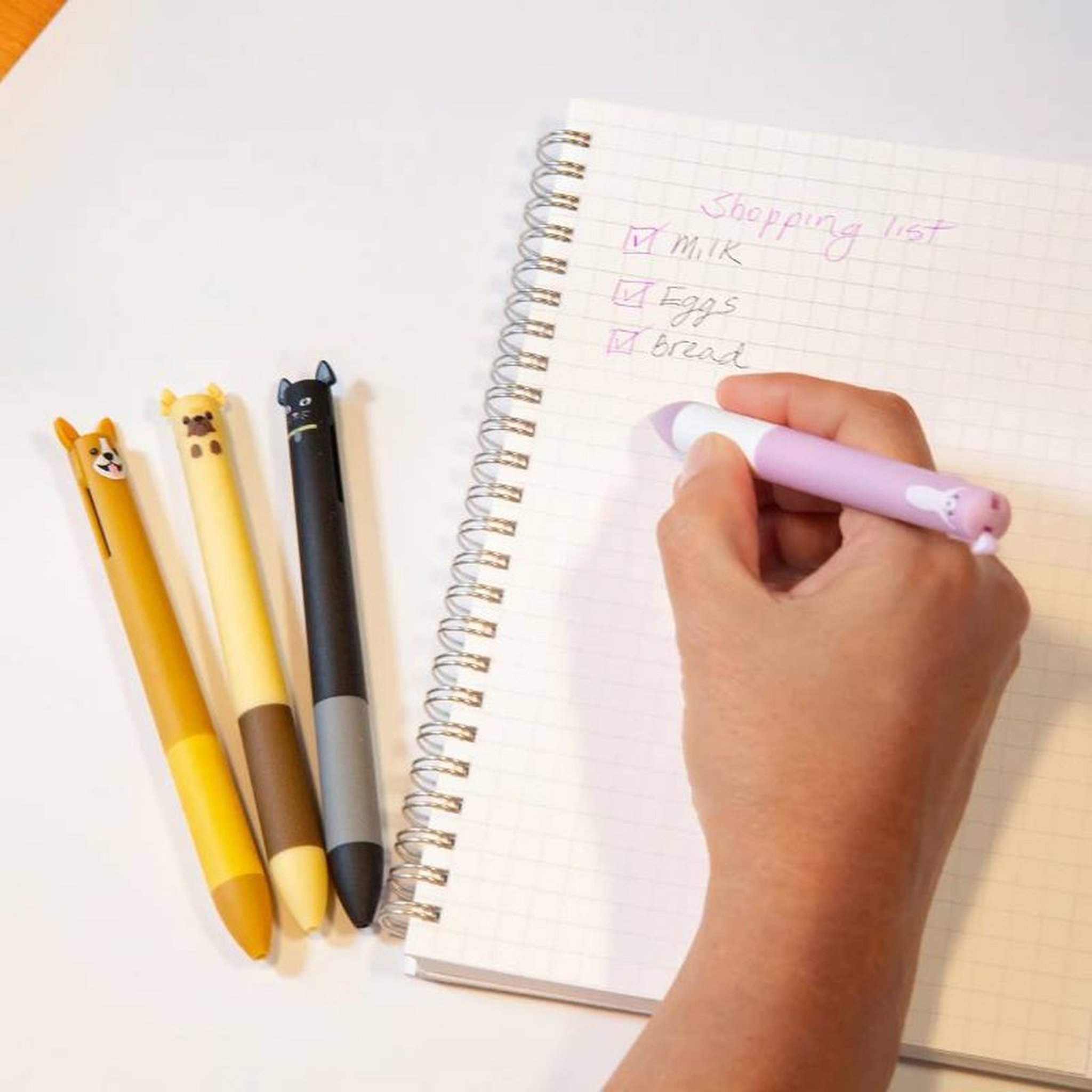 Hand writing a shopping list in the notebook with the cat pen, the other pens beside