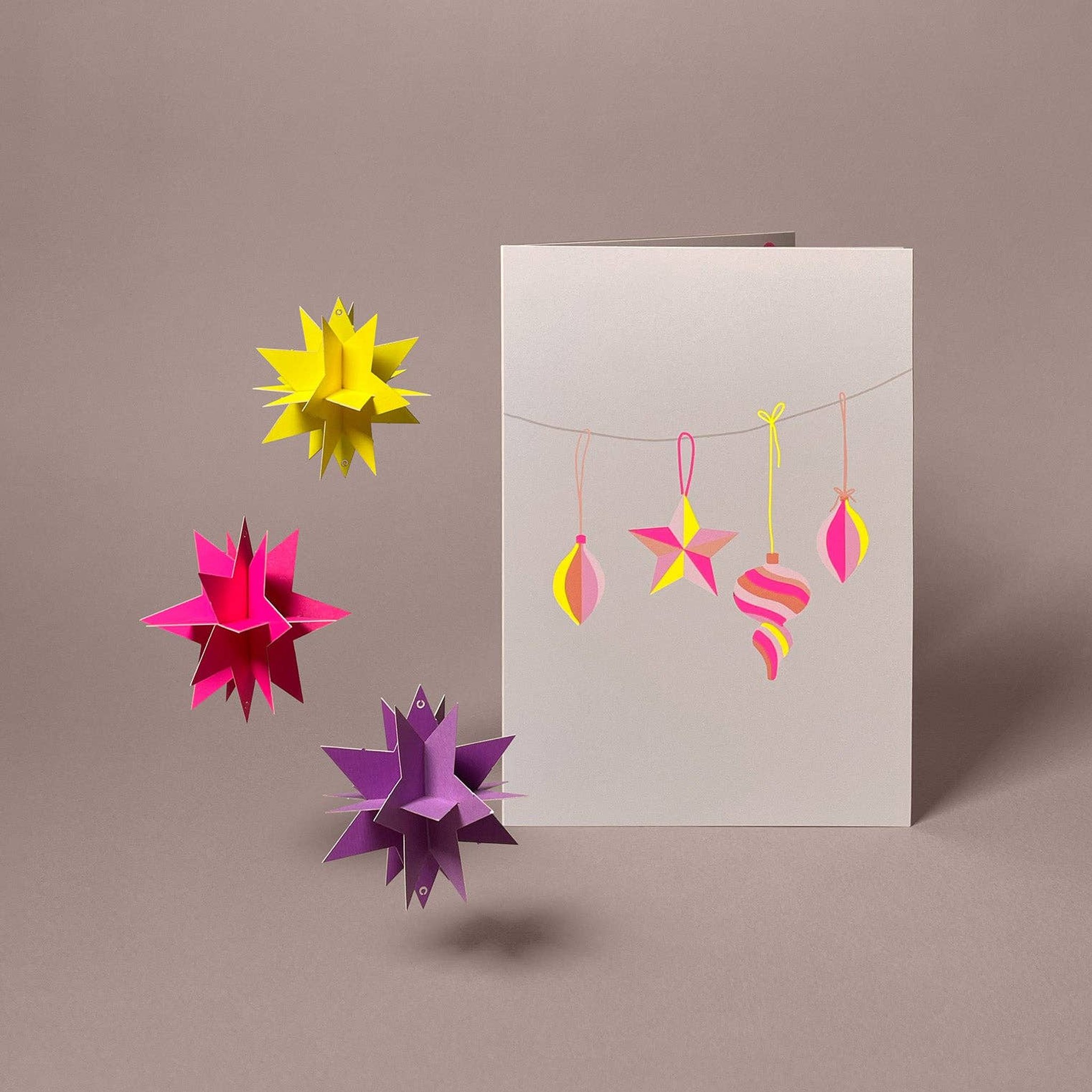 Gefaltetes 3D-Modell drei Sterne in Neonpink, Neongelb und Violett neben der Weihnachtskarte mit Sternengirlande von Faltmeister