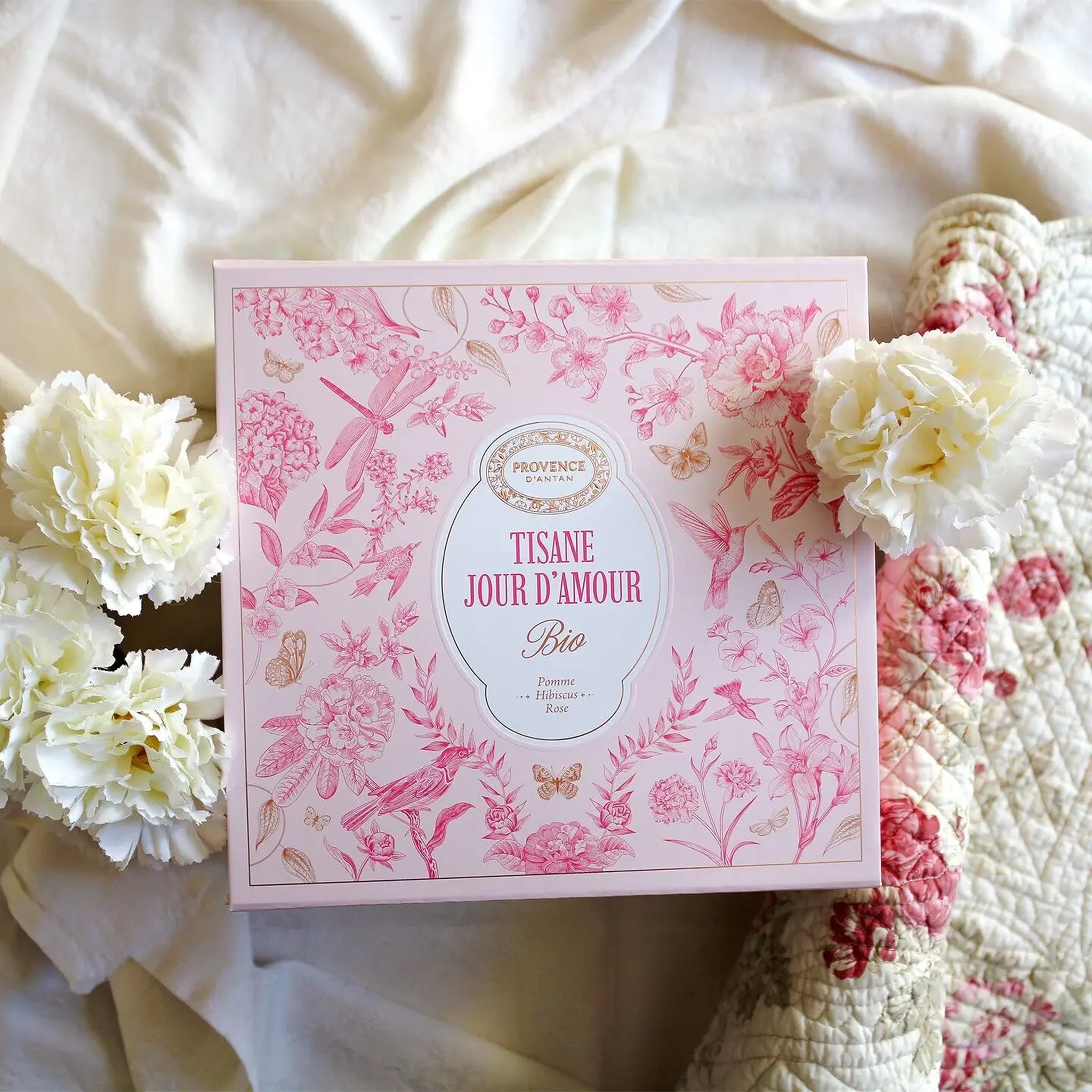 Pink gift box of organic Jour d'Amour herbal tea among white carnations on a patchwork quilt