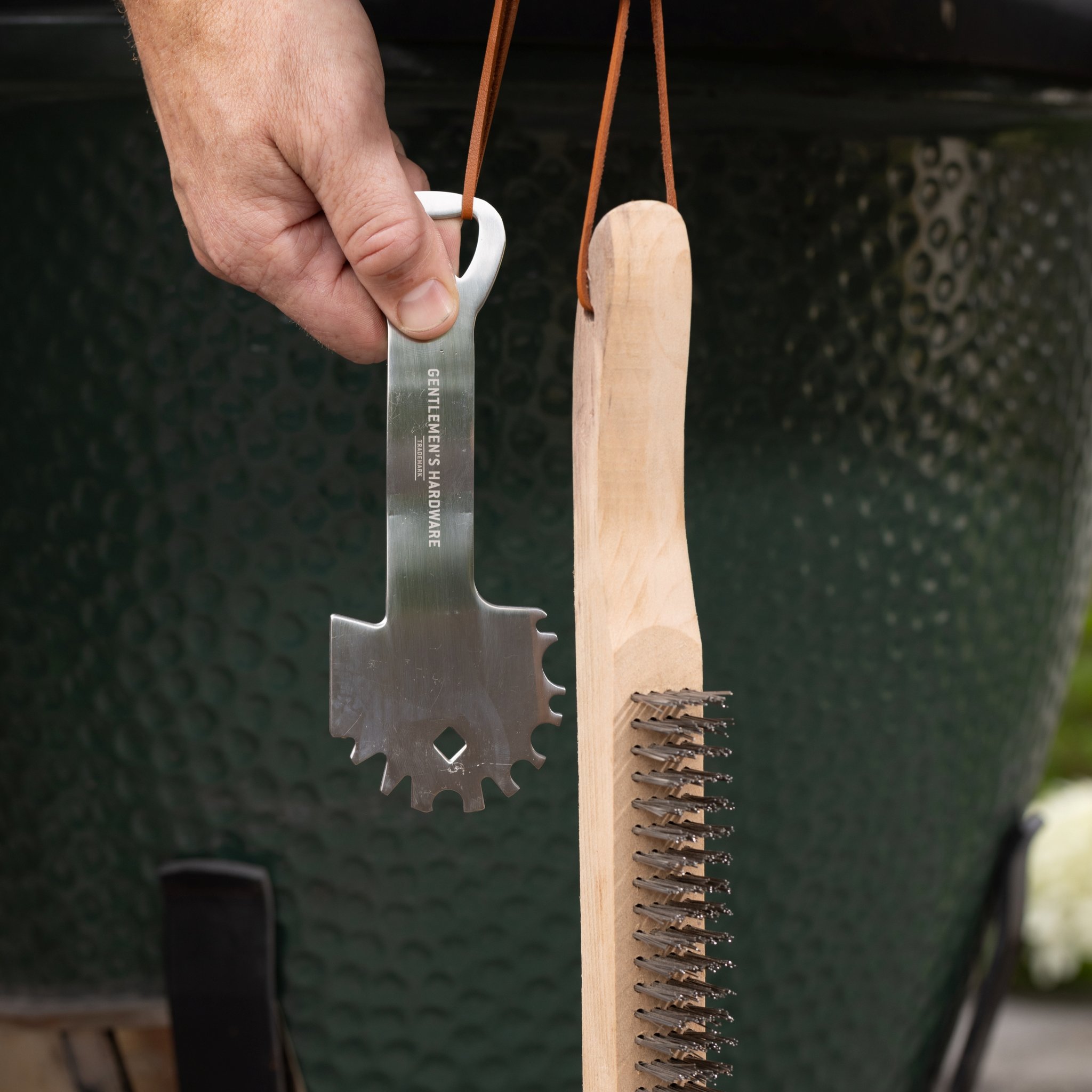 Une main tient le grattoir par le côté décapsuleur, la brosse métallique pend à côté du gril vert, en extérieur