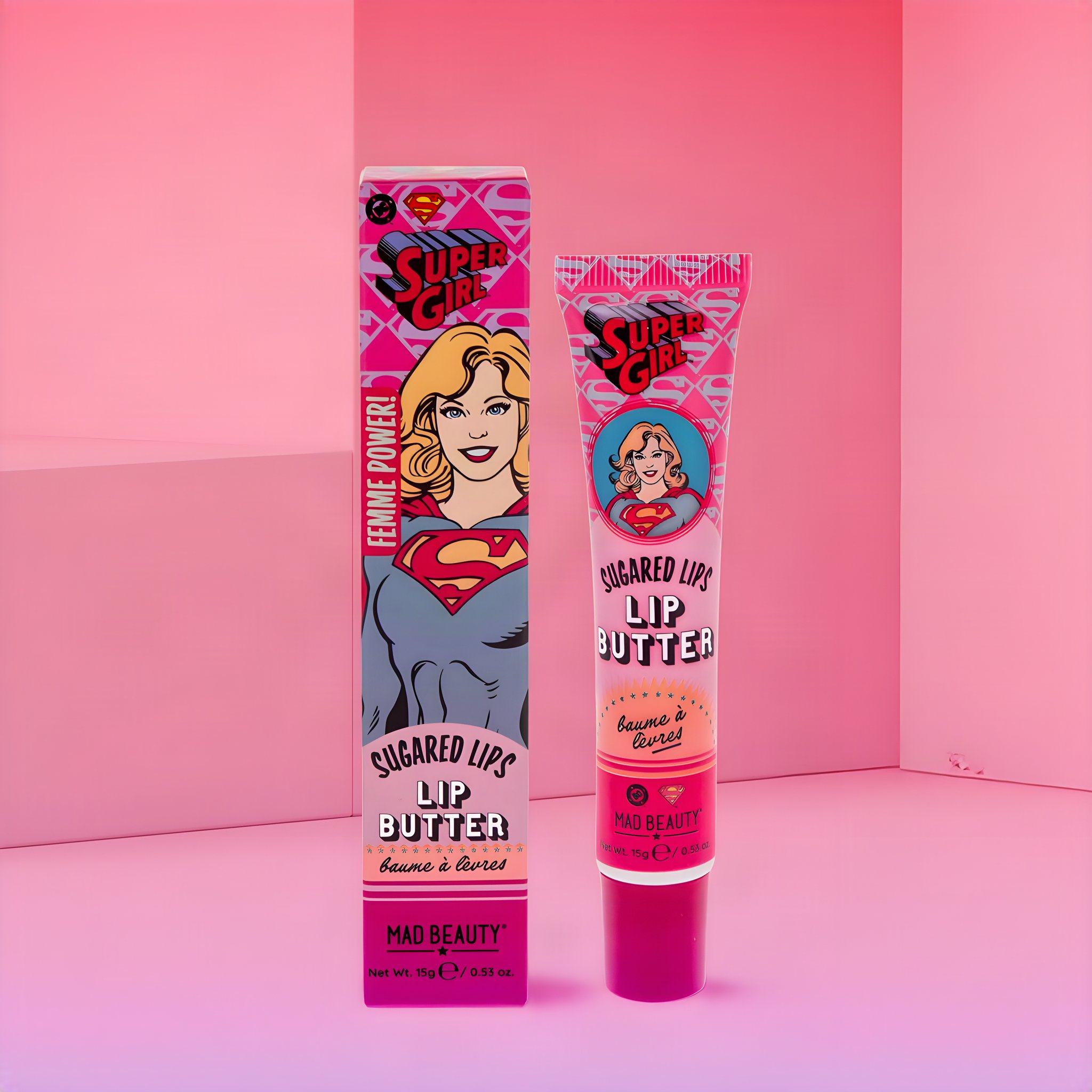 Supergirl Lip Butter: Tube neben der Faltschachtel mit Comic-Supergirl, inszeniert in einem rosa Raum
