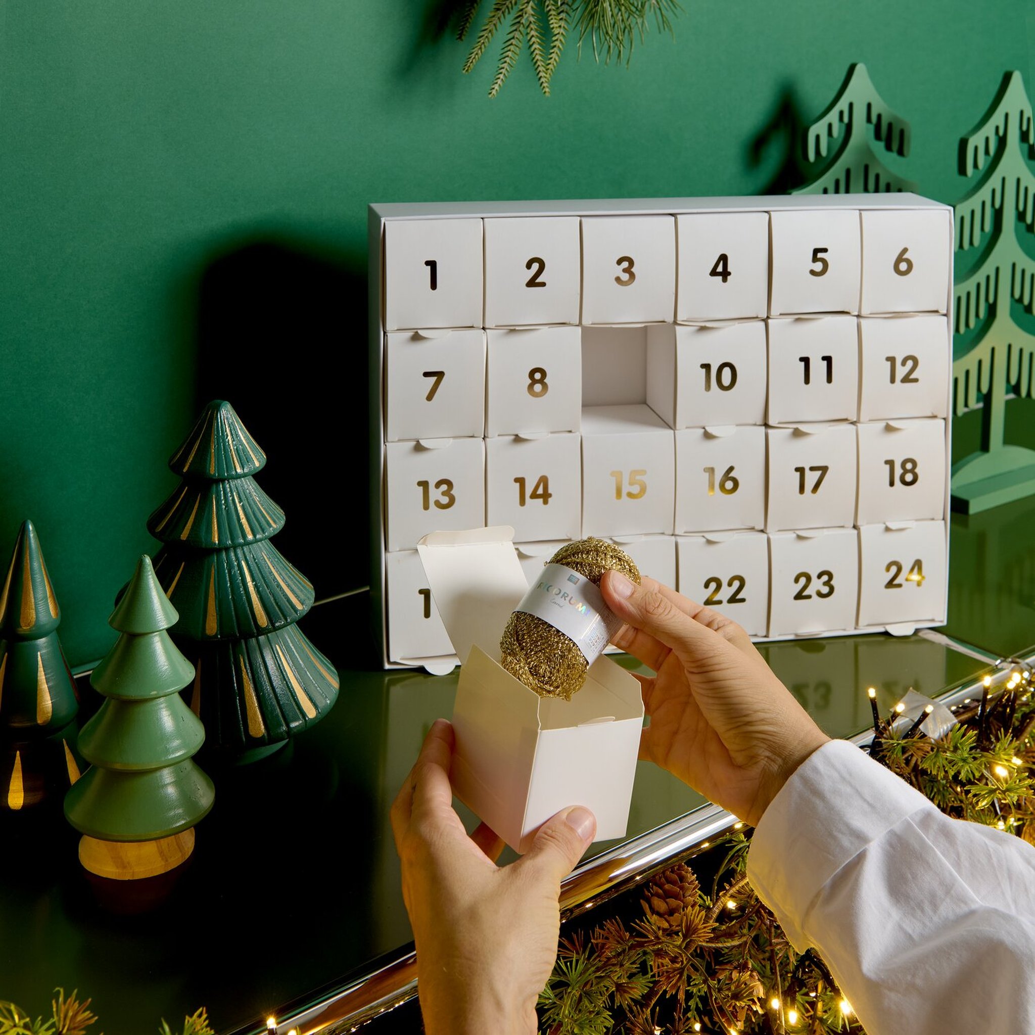 Geöffneter Adventskalender mit 24 nummerierten Schachteln, aus Türchen 15 kommt ein goldenes Glitzerknäuel