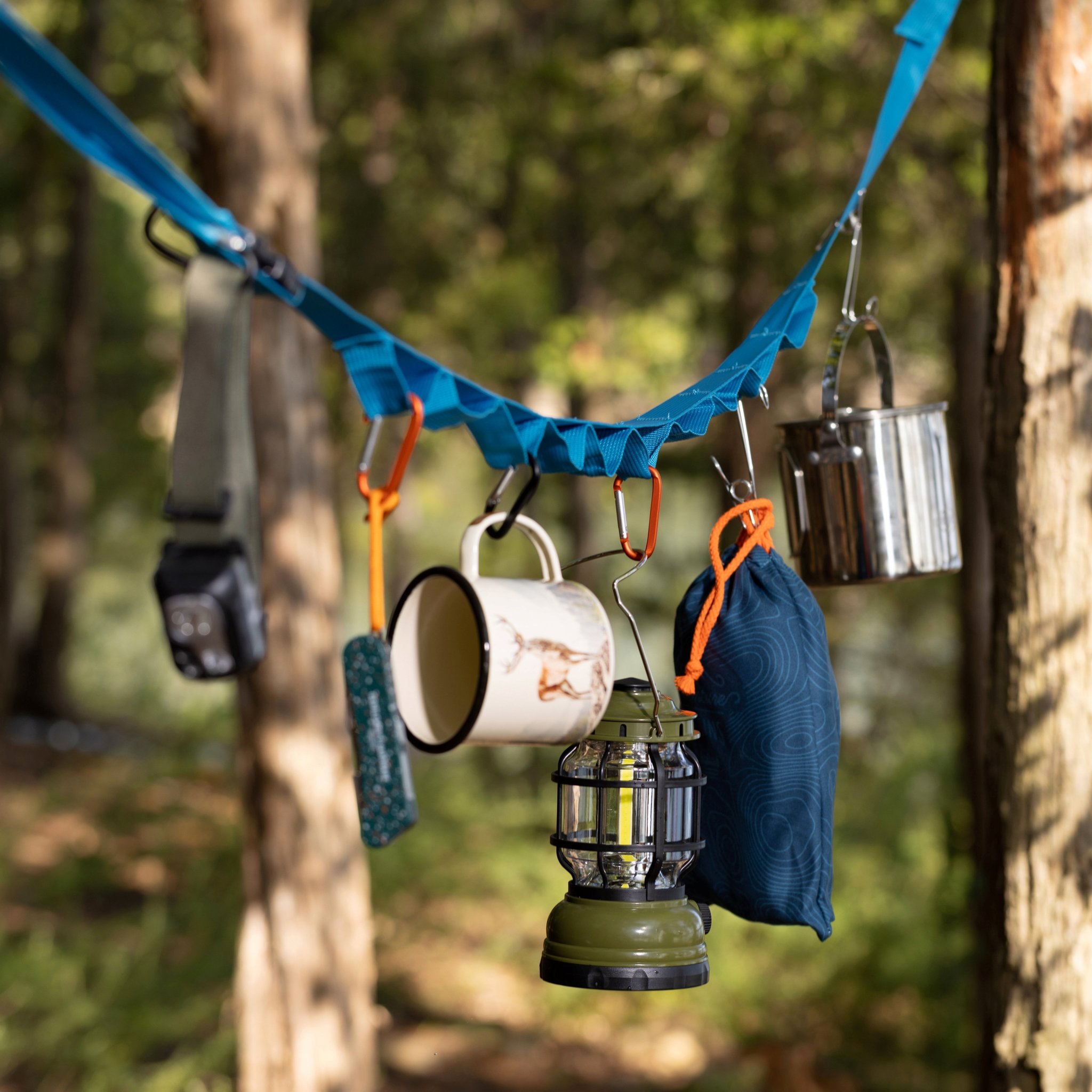 Close view in the woods: lantern and mug on carabiners along the strap, with a pouch and metal pot beside them