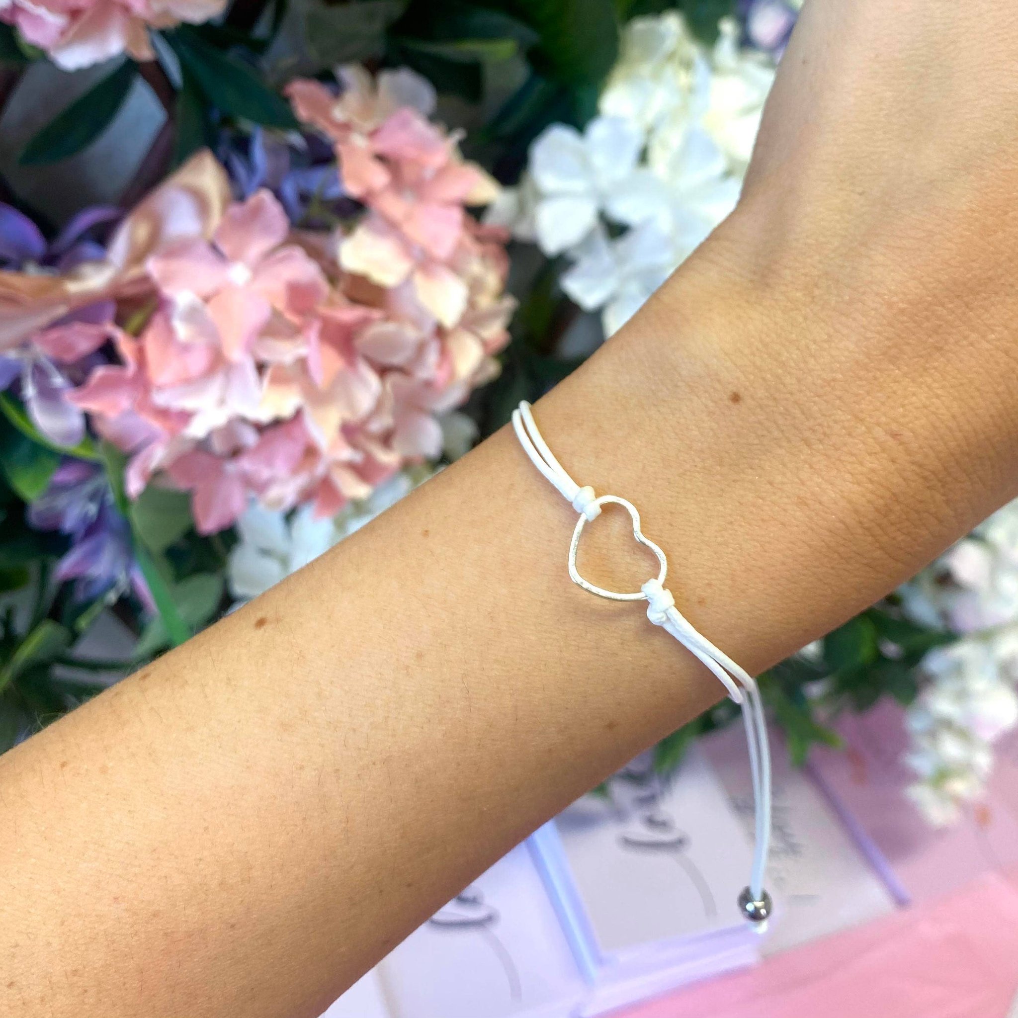 Weisses Fadenarmband mit Herz-Anhänger am Handgelenk vor einem Strauss Hortensien