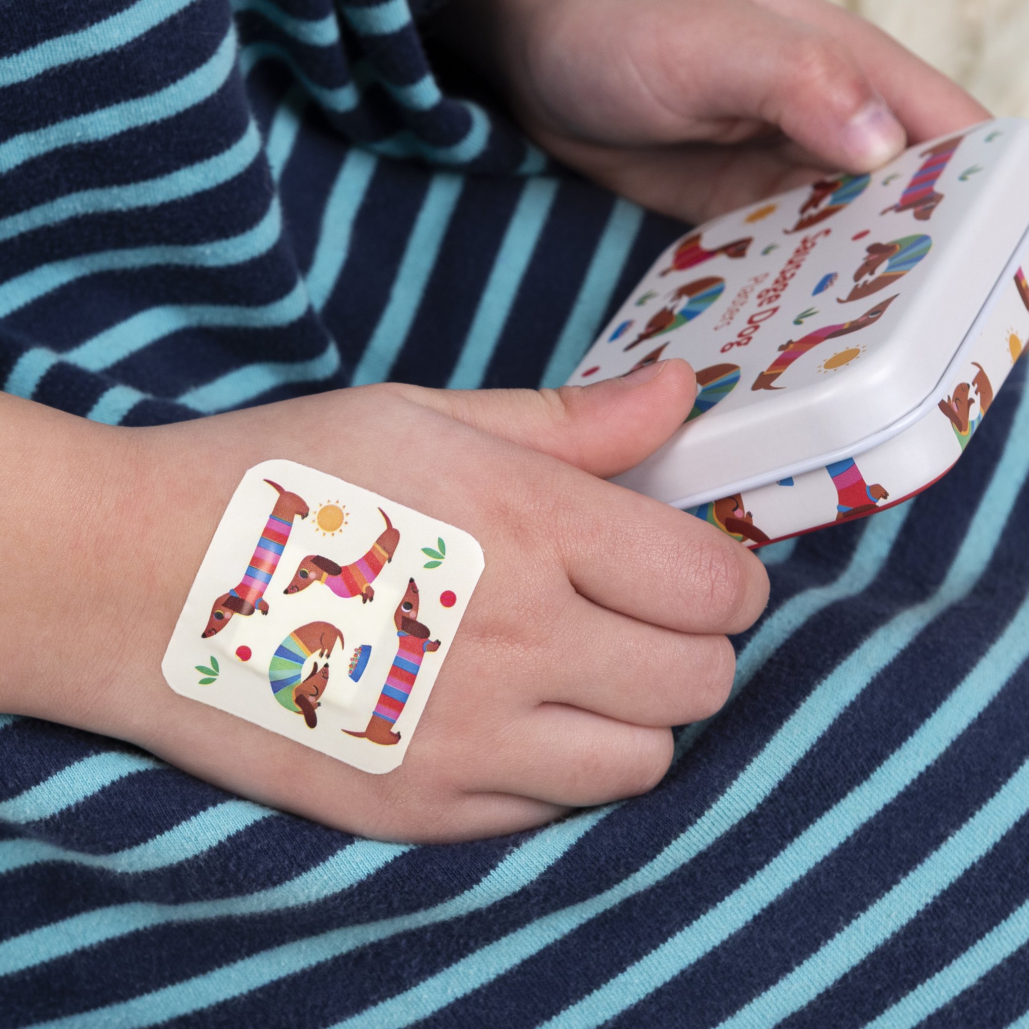 Kinderhand mit Dackel-Pflaster, daneben die Dose der Dackel Kinderpflaster