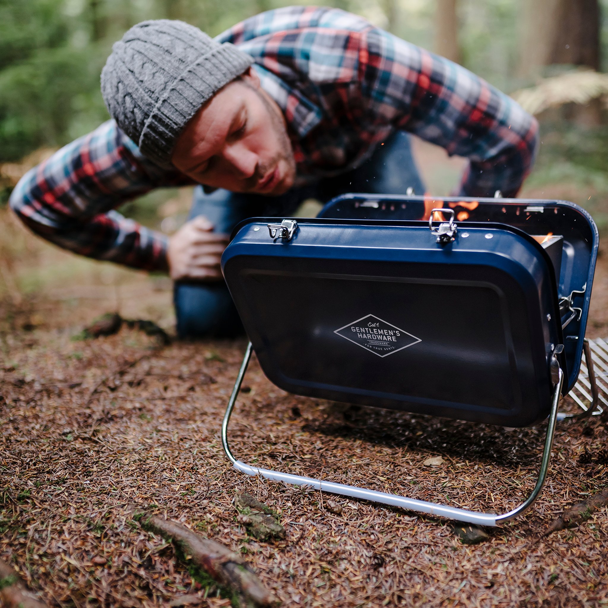 Man in a beanie and checked shirt crouches in the woods, blowing on the embers of the Large Portable BBQ to start the fire