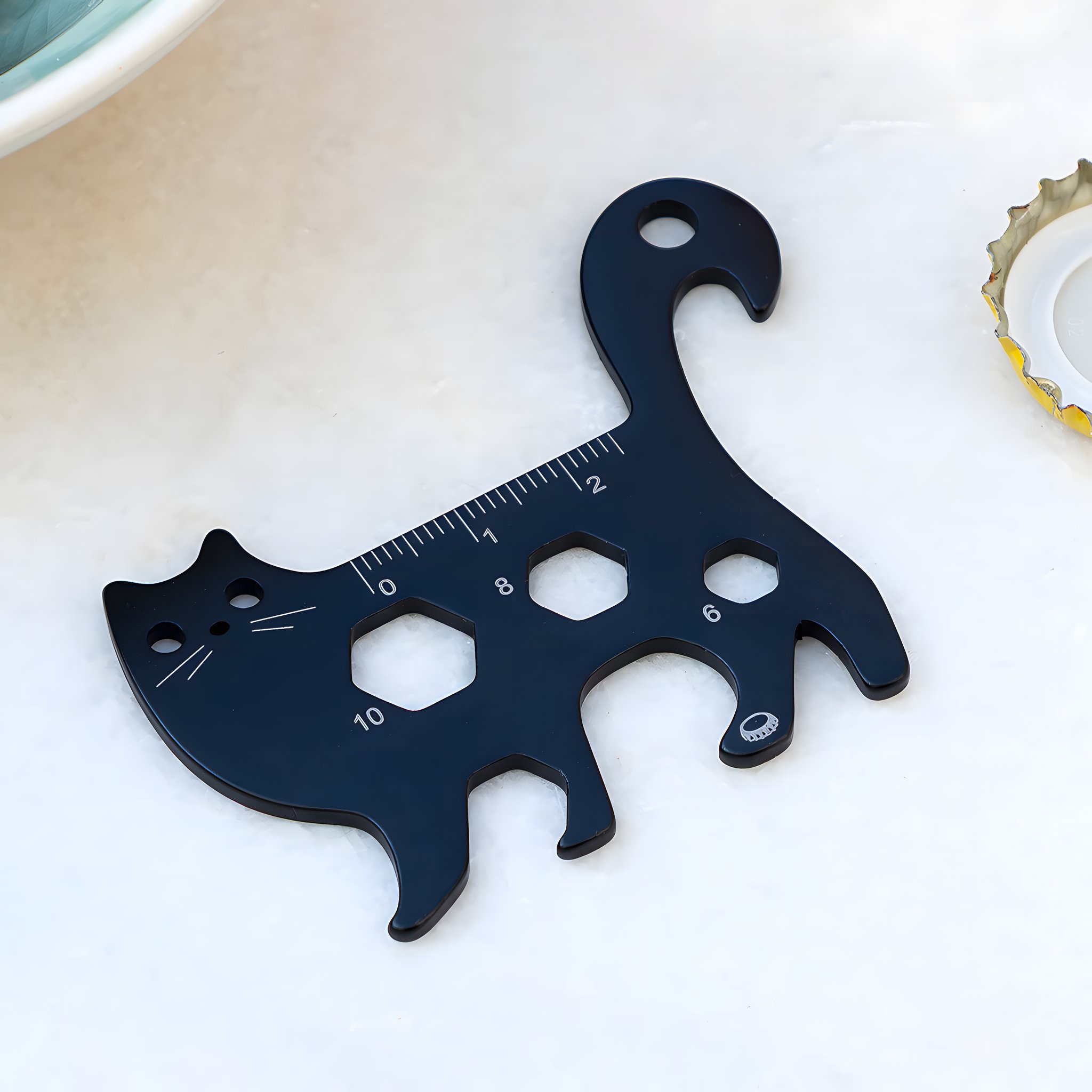 Dunkles Katzen-Multitool mit Lineal-Skala neben einem Kronkorken auf hellem Tisch