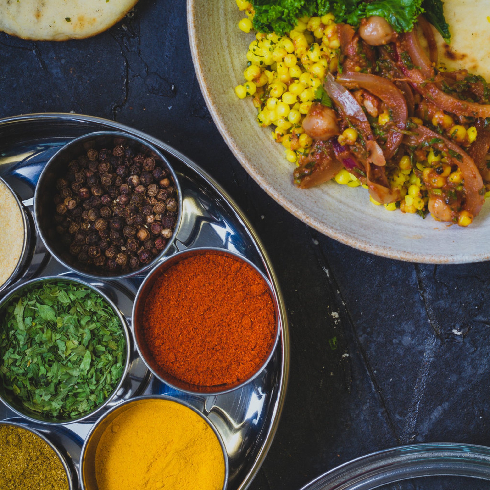 Masala Dabba mit Gewürzen neben Curry und Naan auf dunklem Untergrund