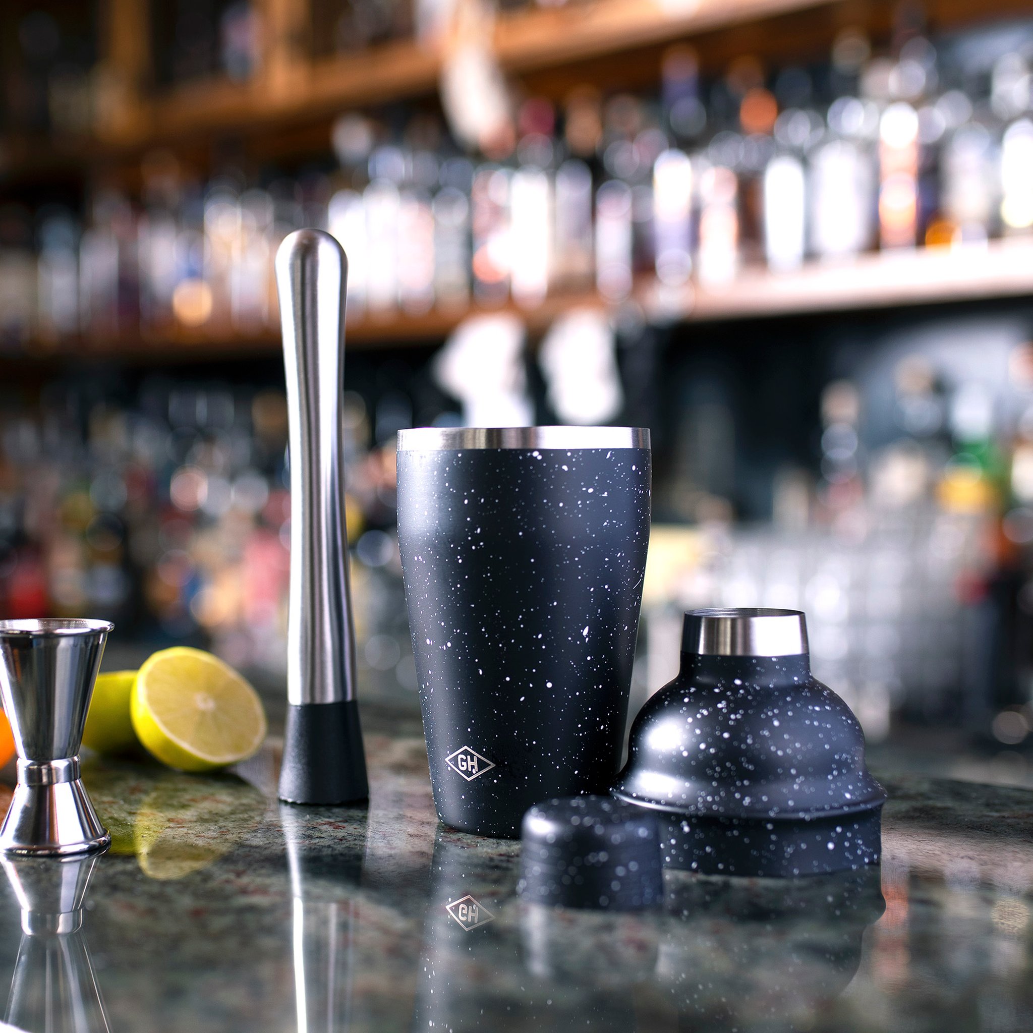 Mixology kit on a stone bar counter with a halved lemon, bottles on shelves glowing out of focus behind