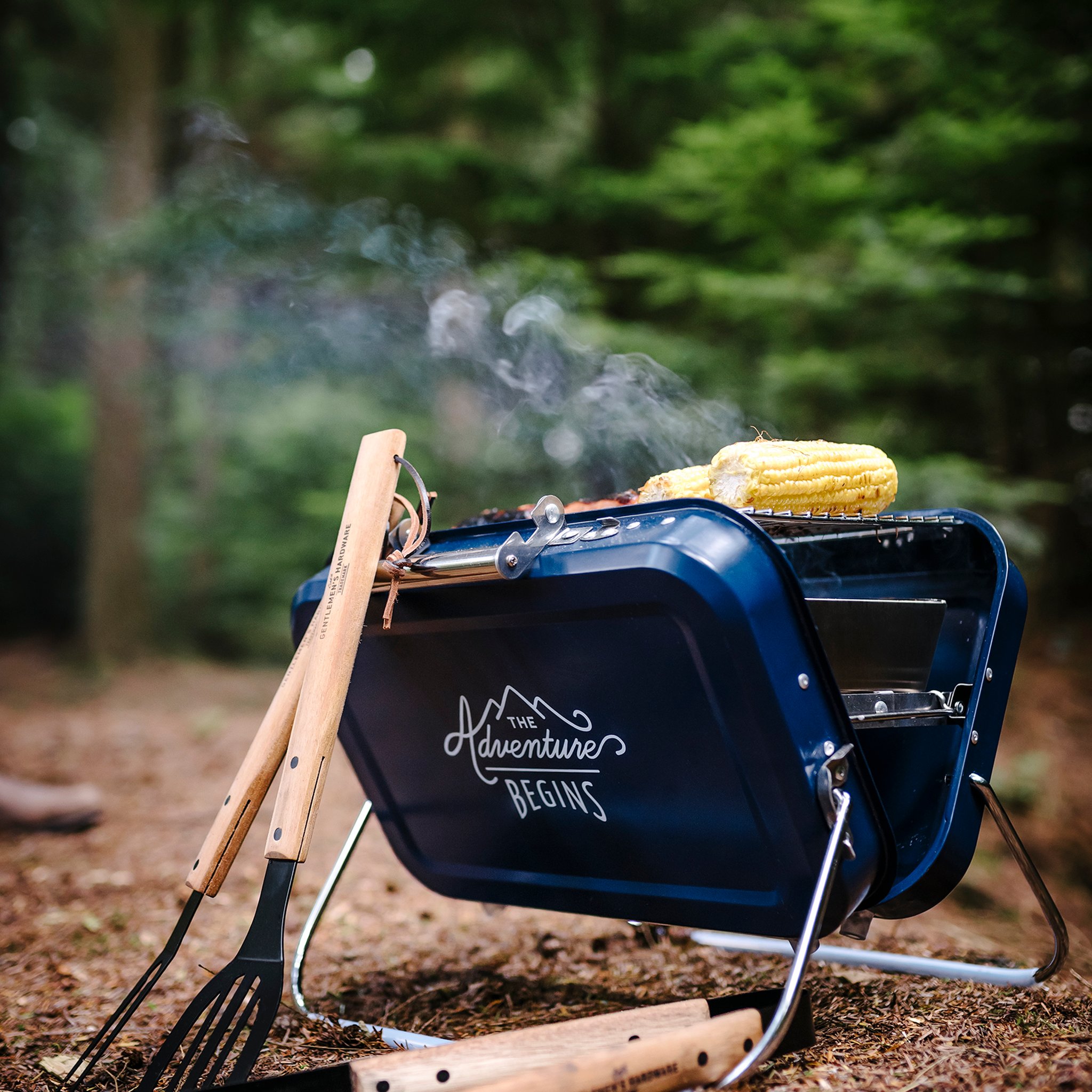 Large Portable BBQ in a forest with corn cobs on the grill, rising smoke and barbecue tools leaning against it