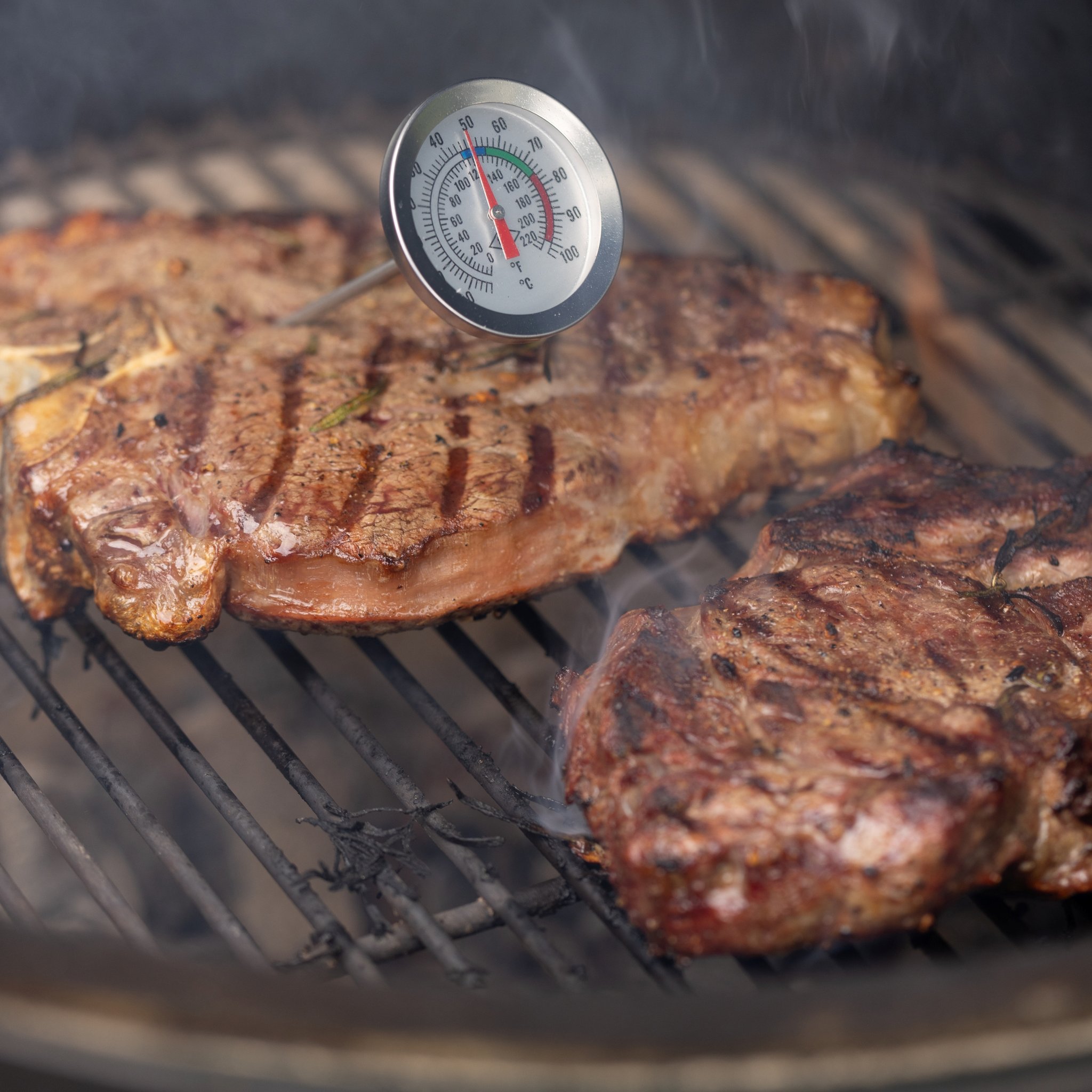 Meat thermometer inserted in a seared steak with grill marks on a smoking grate, a second cut beside it