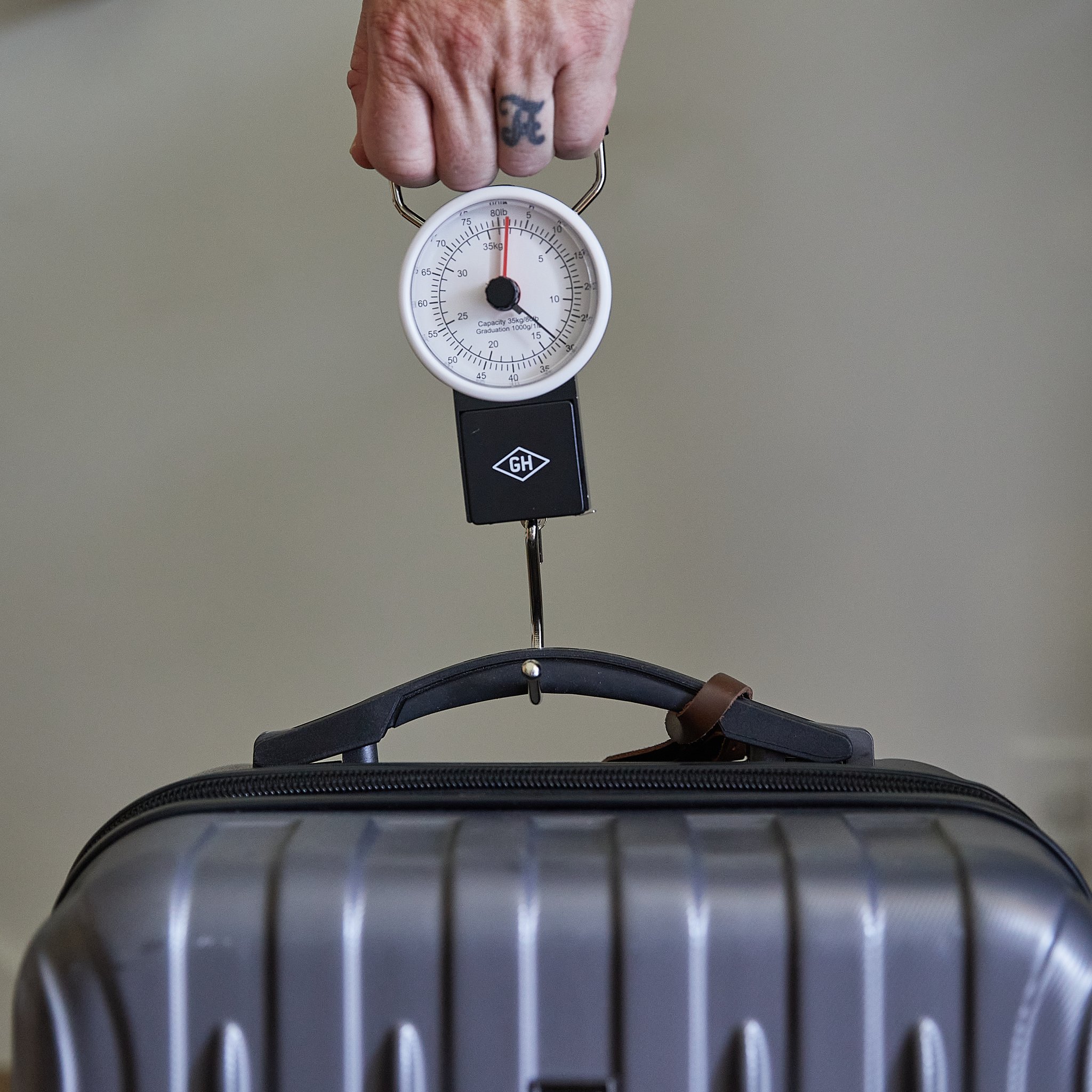 A hand lifts a grey hard-shell suitcase by its handle using the Travel Scale hooked on to weigh it