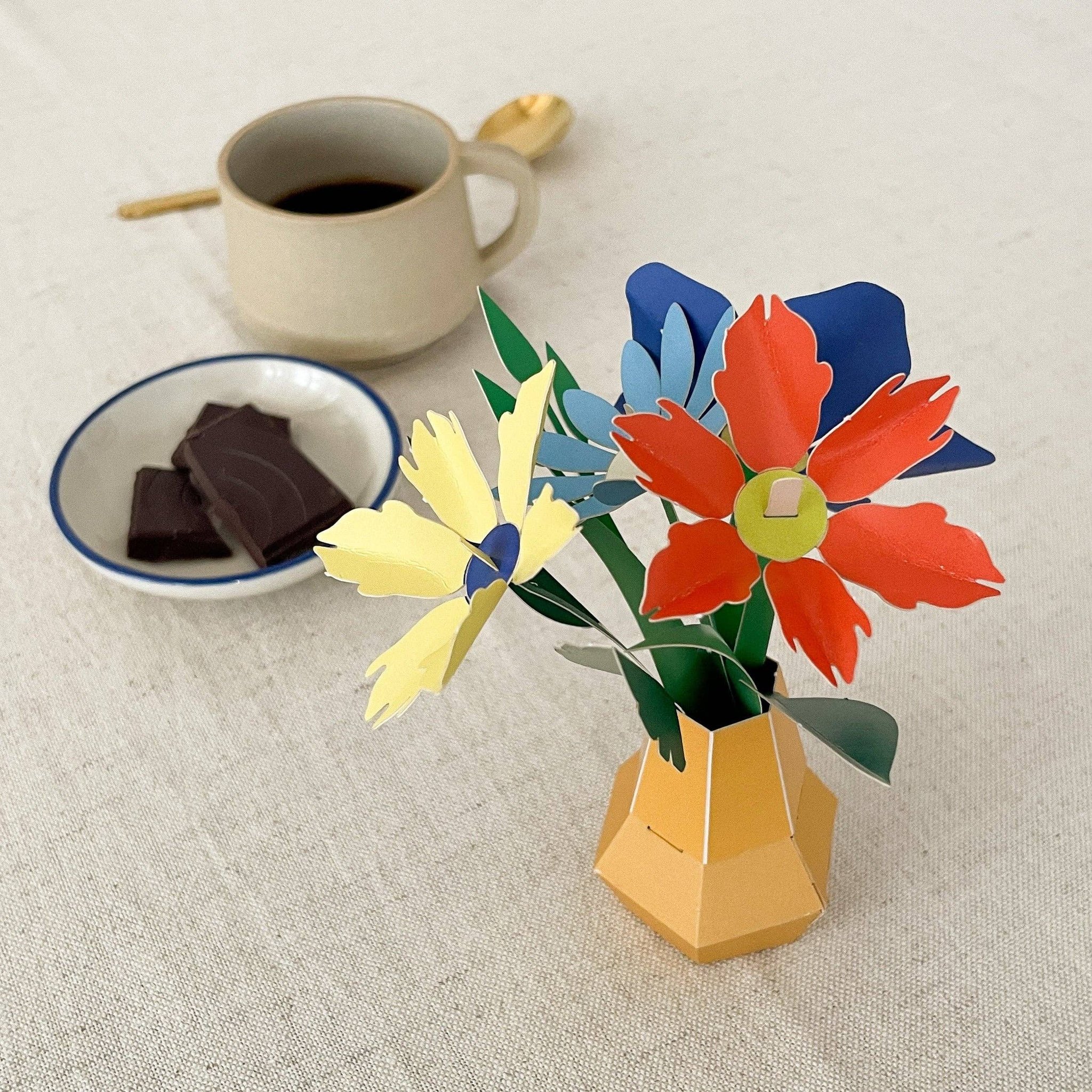 Papier-Blumenstrauss in gelber Vase neben Kaffeetasse und Schokolade