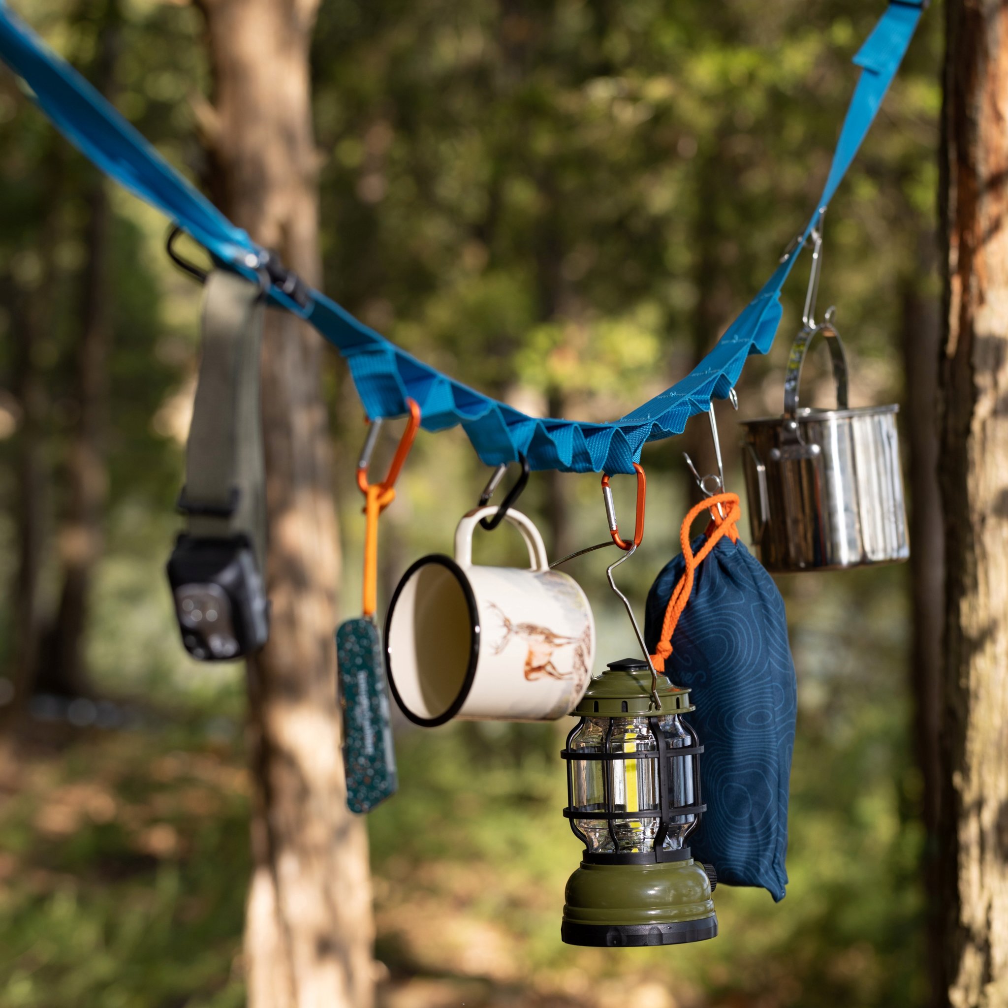 Strap strung between forest trees holding a headtorch, enamel mug, lantern and pot, the Hanging tent tidy in use